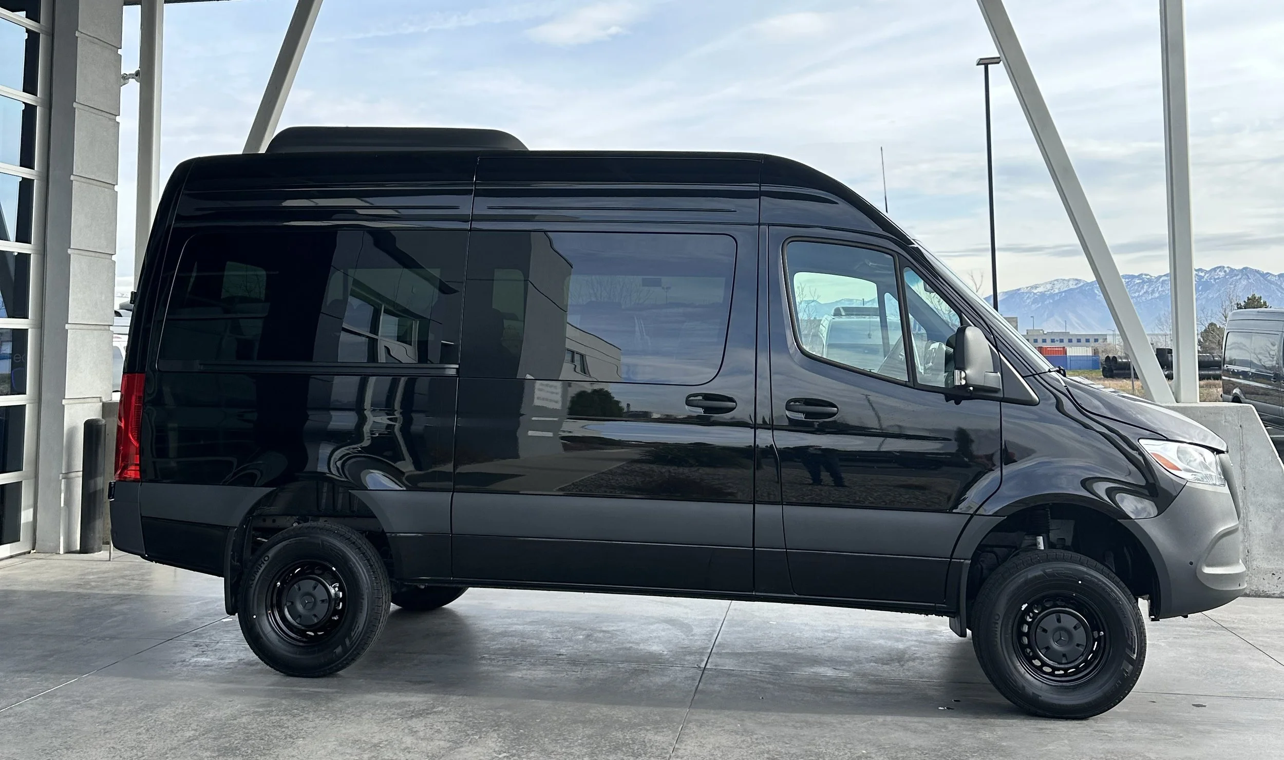 A black cargo van with missing wheels parked on a concrete surface under a partially covered structure, with mountains and a partly cloudy sky in the background.