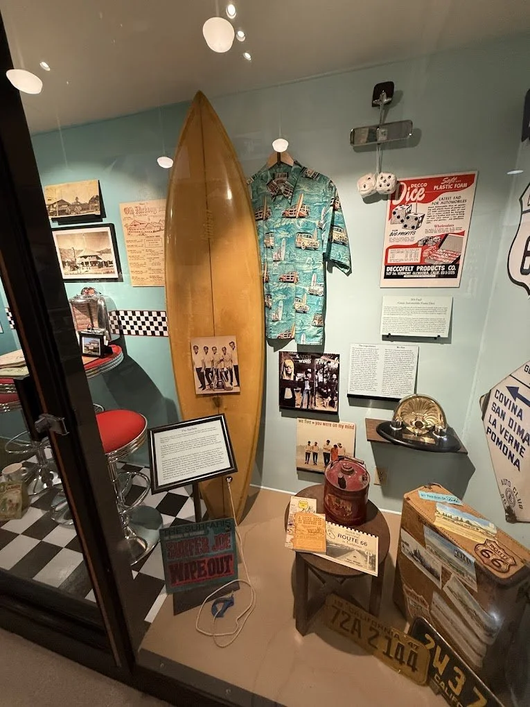 Display case at the Glendora Museum featuring a vintage surfboard, a Hawaiian shirt, model cars, old roadside signs, a jukebox, vintage cameras, and travel memorabilia including Route 66 and license plates.