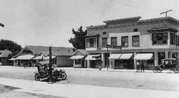 A historic black and white photograph of downtown glendora with storefronts and awnings, a vintage car, and a horse-drawn carriage.