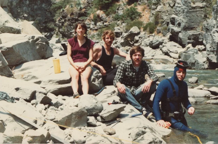 Four people sitting on rocks beside a river in the San Gabriel Mountains, with one person wearing a wetsuit and holding a rope.