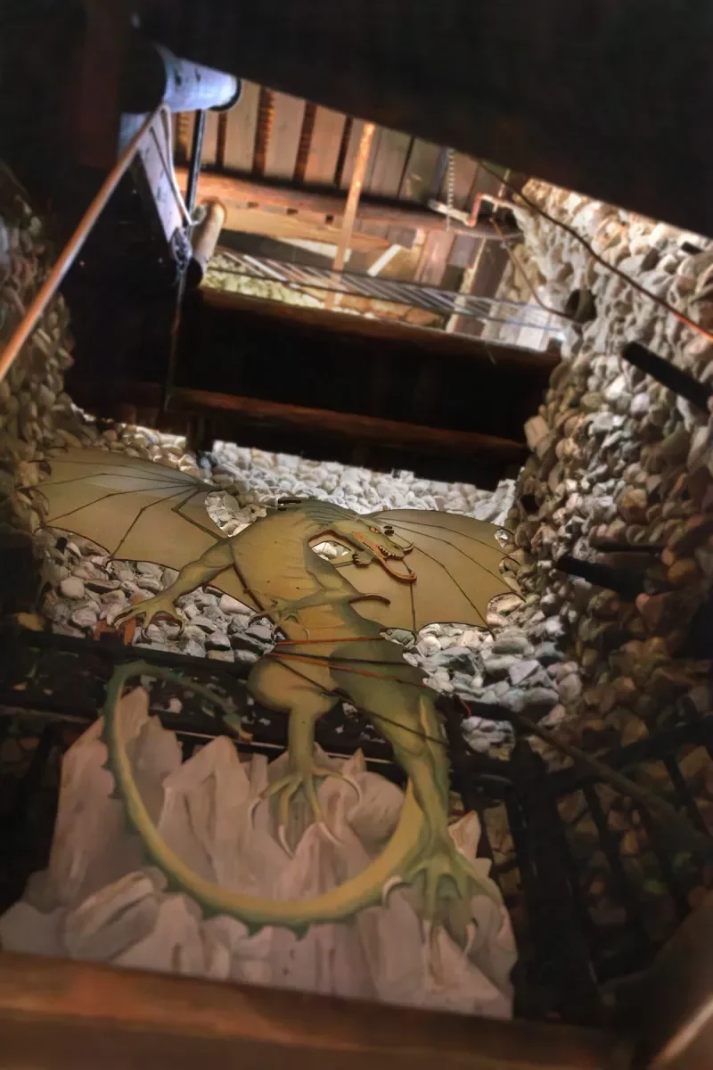 Visitors to the rebel castle enter through a tunnel. If they look up they will see this painting of a green flying dragon with large wings, perched on a rocky base, surrounded by stone walls and exposed wooden beams.