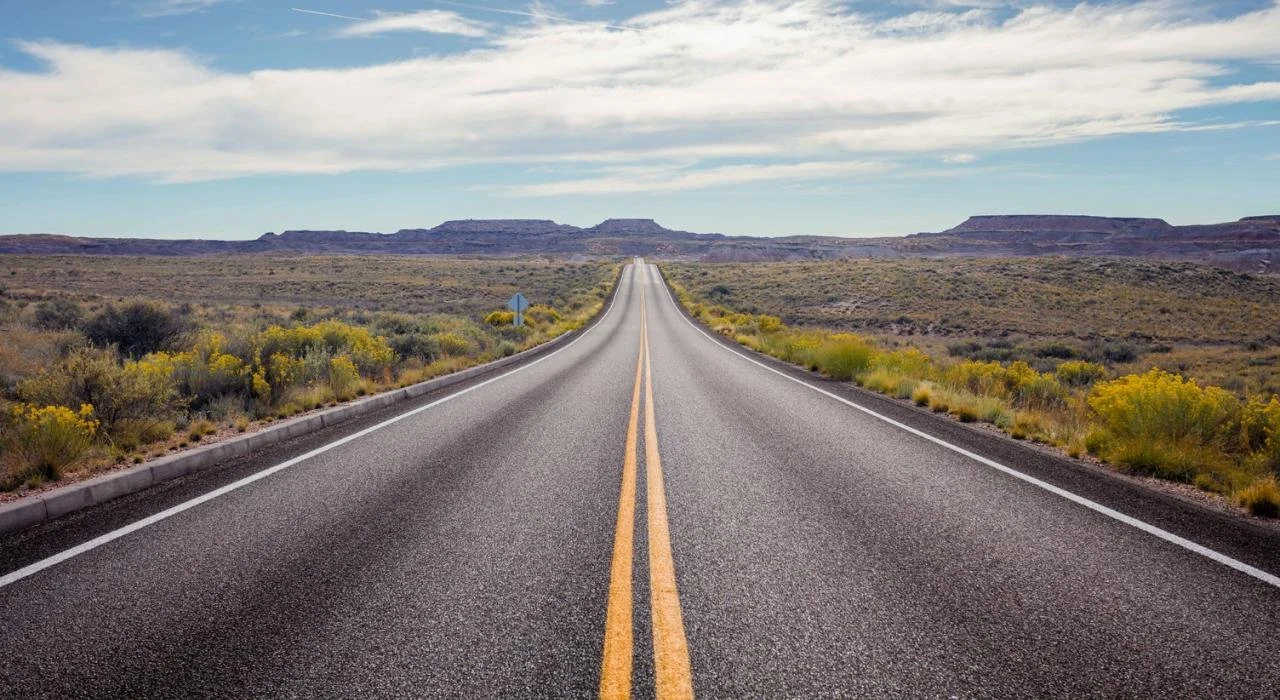 A straight stretch of Route 66 extending into the distance through a desert landscape with sparse shrubs and distant mesas under a partly cloudy sky.