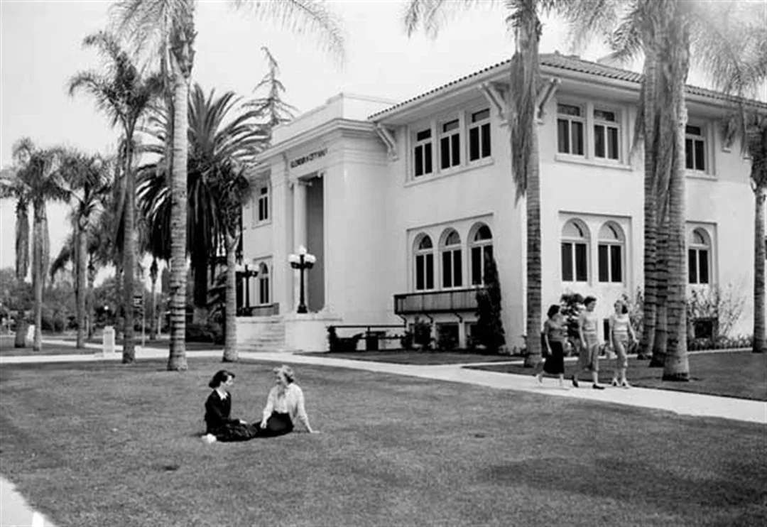 1950s photo of two women sitting on the grass in front of Glendora City Hall with palm trees around them, and three people walking on a sidewalk nearby.