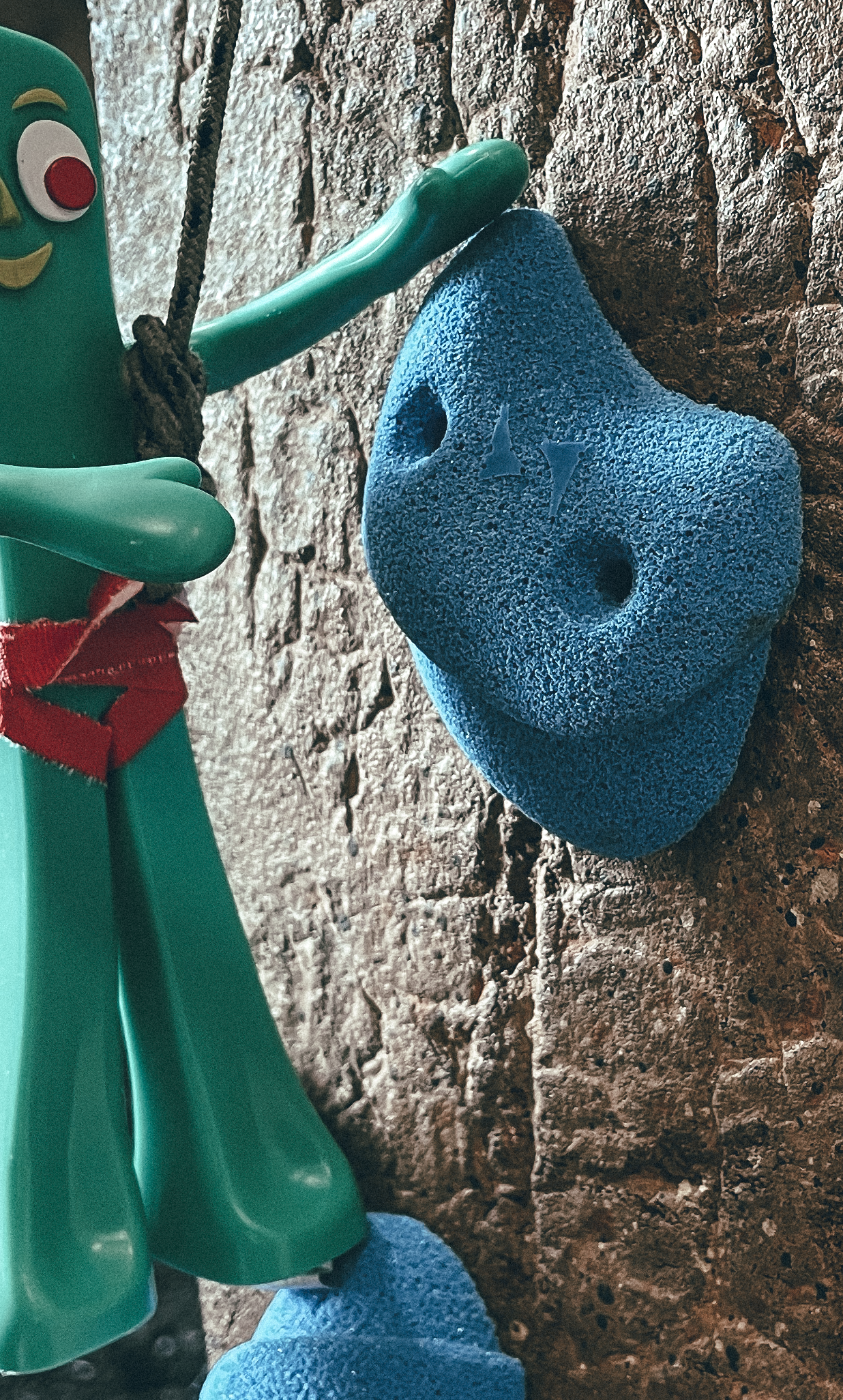 A close-up view of a green Gumby figuring attached to a textured rock climbing wall.