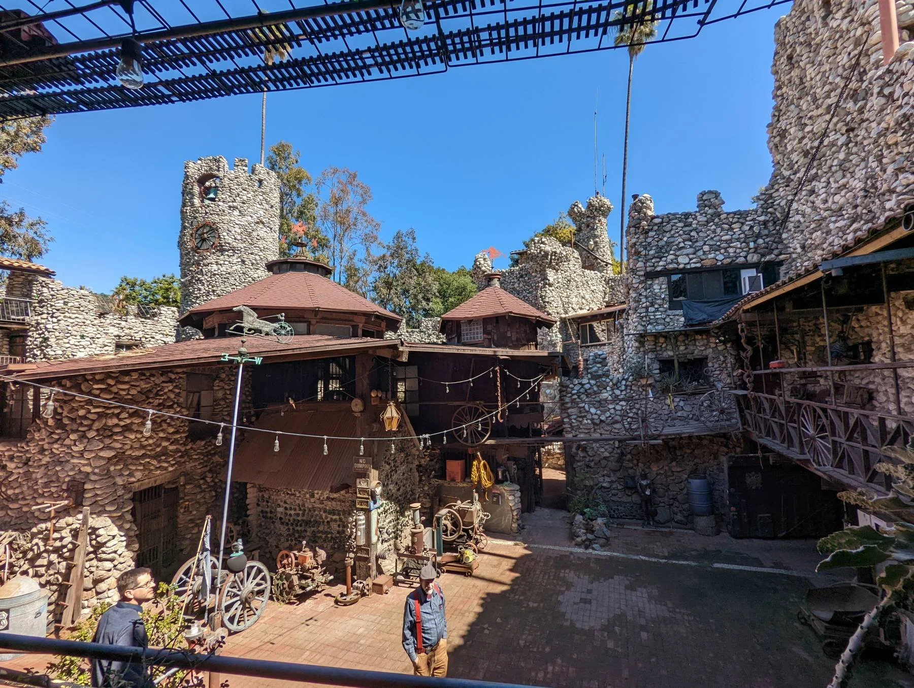 An interior photo of the Rubel Castle courtyard with stone towers, rustic wooden buildings, and vintage artifacts, under a clear blue sky. There are a few people walking and standing in the area.