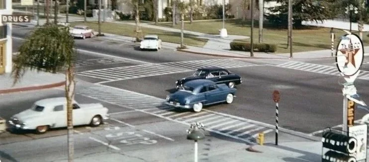 Historic color photo of the intersection of Michigan Avenue (now Glendora Avenue) and Foothill Boulevard Street with cars driving and stopped at a crosswalk, trees lining the sidewalk, and a period Texaco gas station sign.