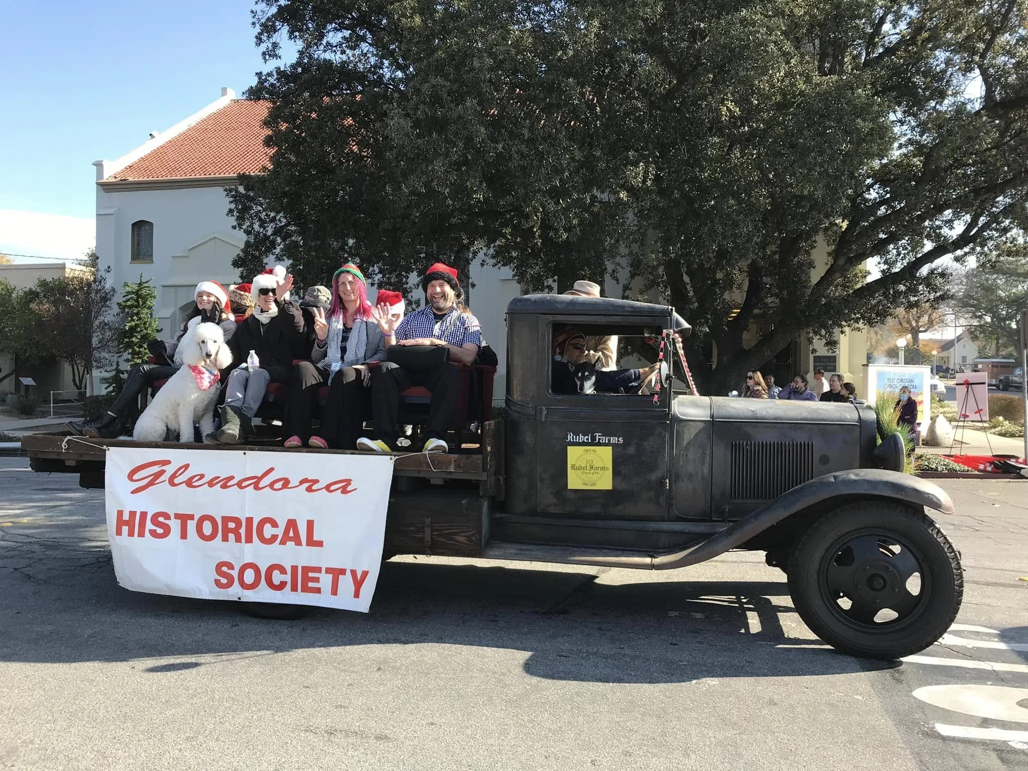 A photo from the Glendora Christmas Parade. An antique truck with people sitting in the truck bed and a banner on the side reading 'Glendora Historical Society' is carrying people and dogs dressed in Christmas attire.