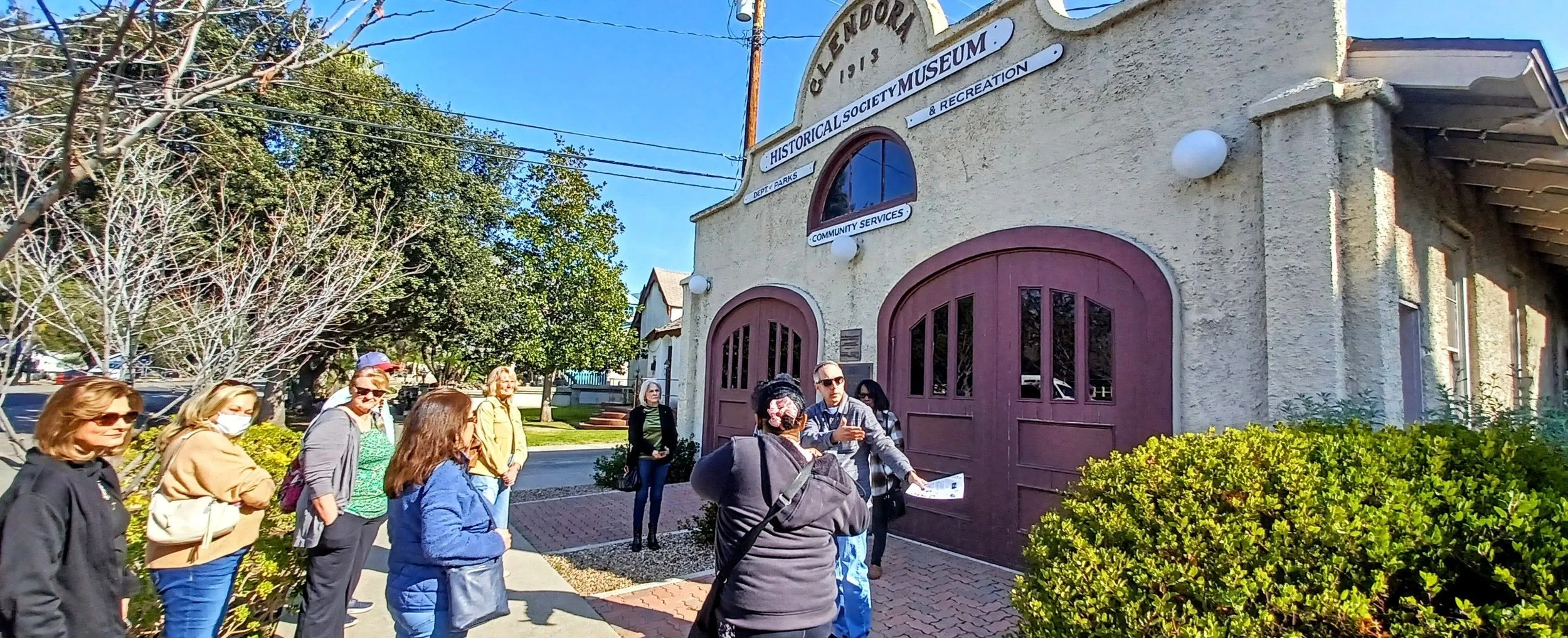 Group of people on a tour standing outside the Glendora Historical Society Museum, a beige building with traditional carriage-style garage doors, on a sunny day.