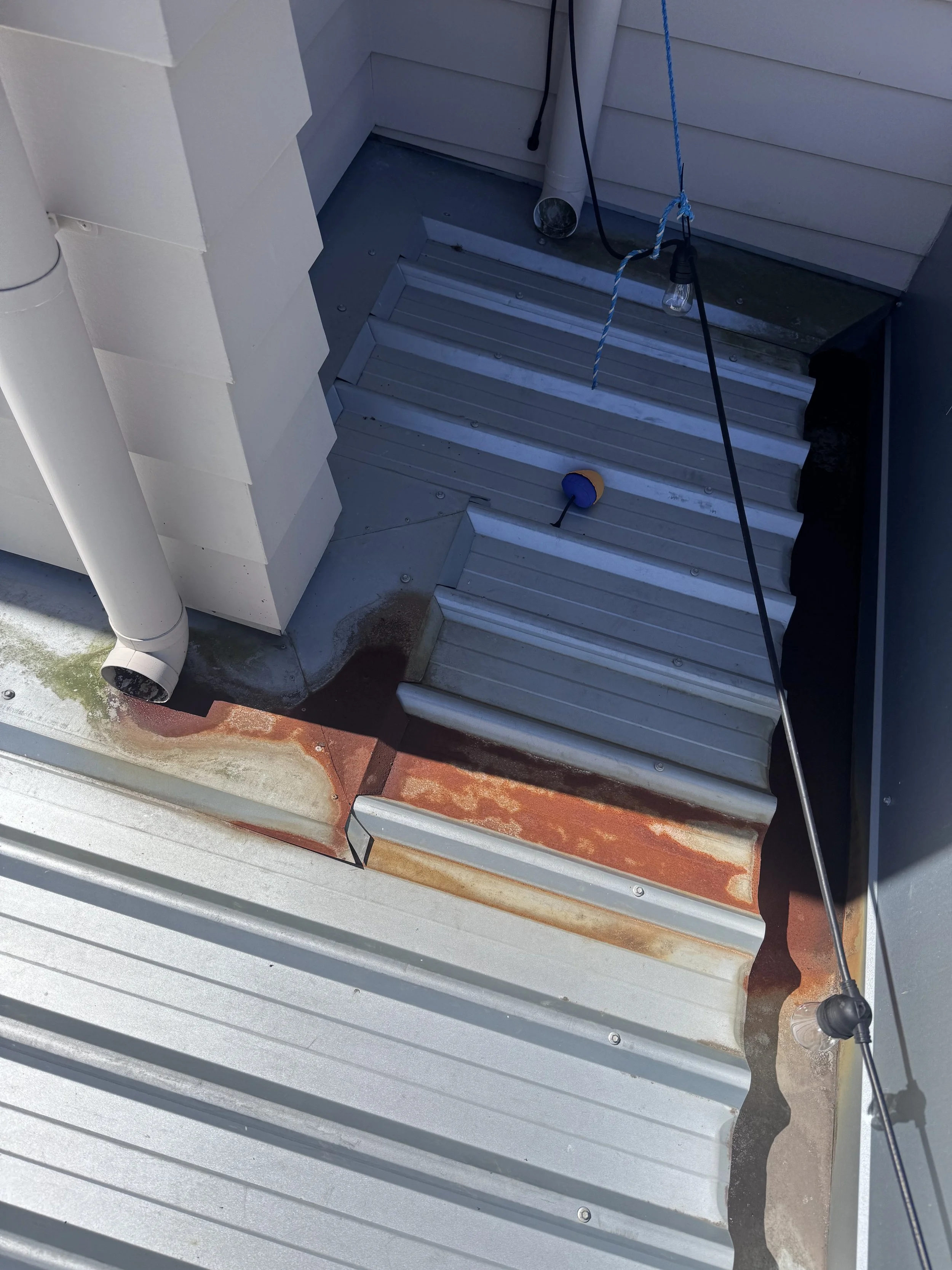 Rusted and corroded section of a metal roof with a downspout and electrical wires hanging down.