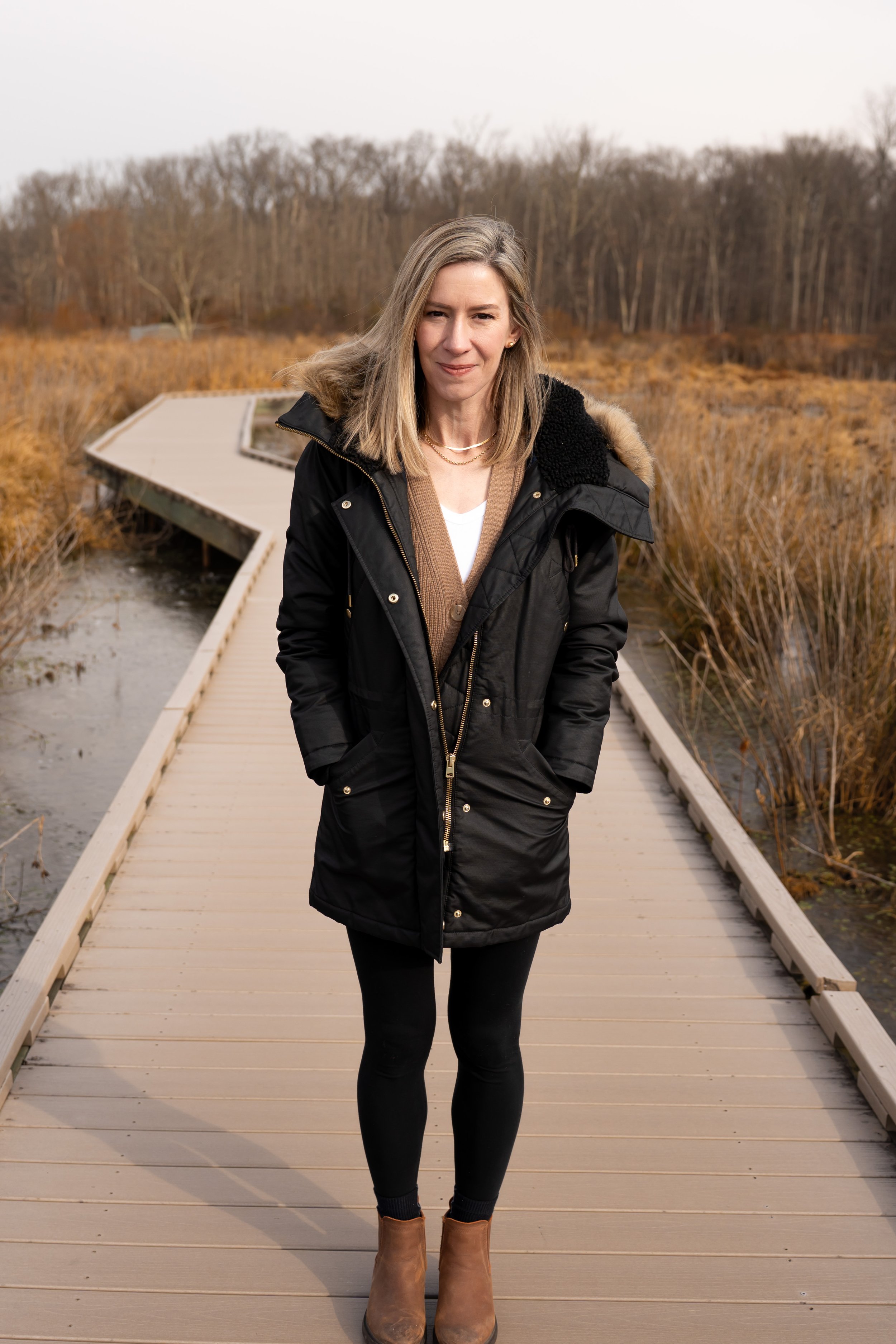 A woman standing on a wooden boardwalk outdoors, surrounded by autumnal bushes and trees, wearing a black coat and brown ankle boots.