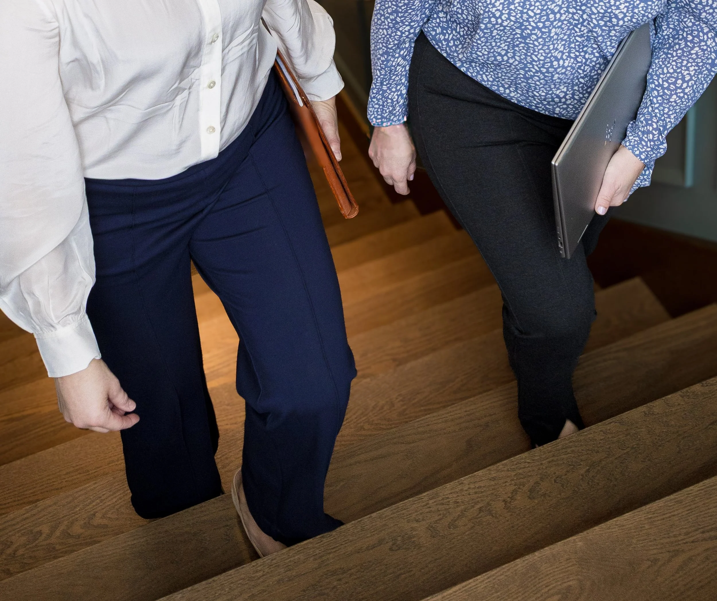 Two women going up wooden stairs, one holding a laptop and the other holding a folder.
