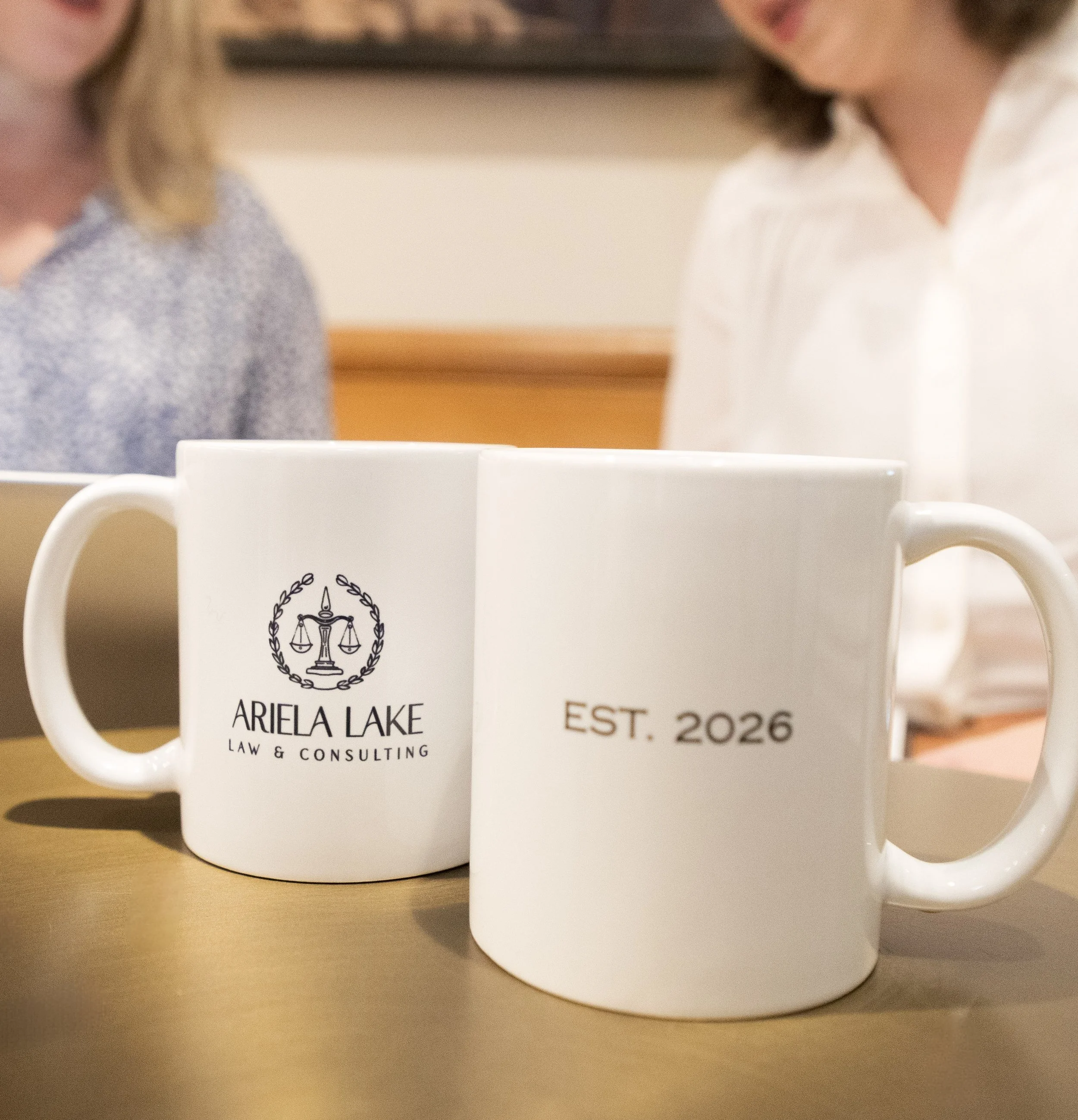 Two white coffee mugs on a wooden table, one with the logo and text 'ARIELA LAKE LAW & CONSULTING' and the other with 'EST. 2026', with blurred people in the background.