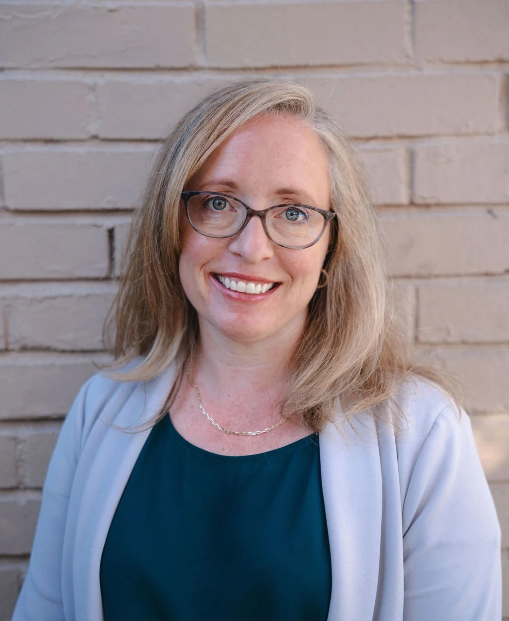 A woman with blonde hair, glasses, and a friendly smile standing in front of a brick wall.