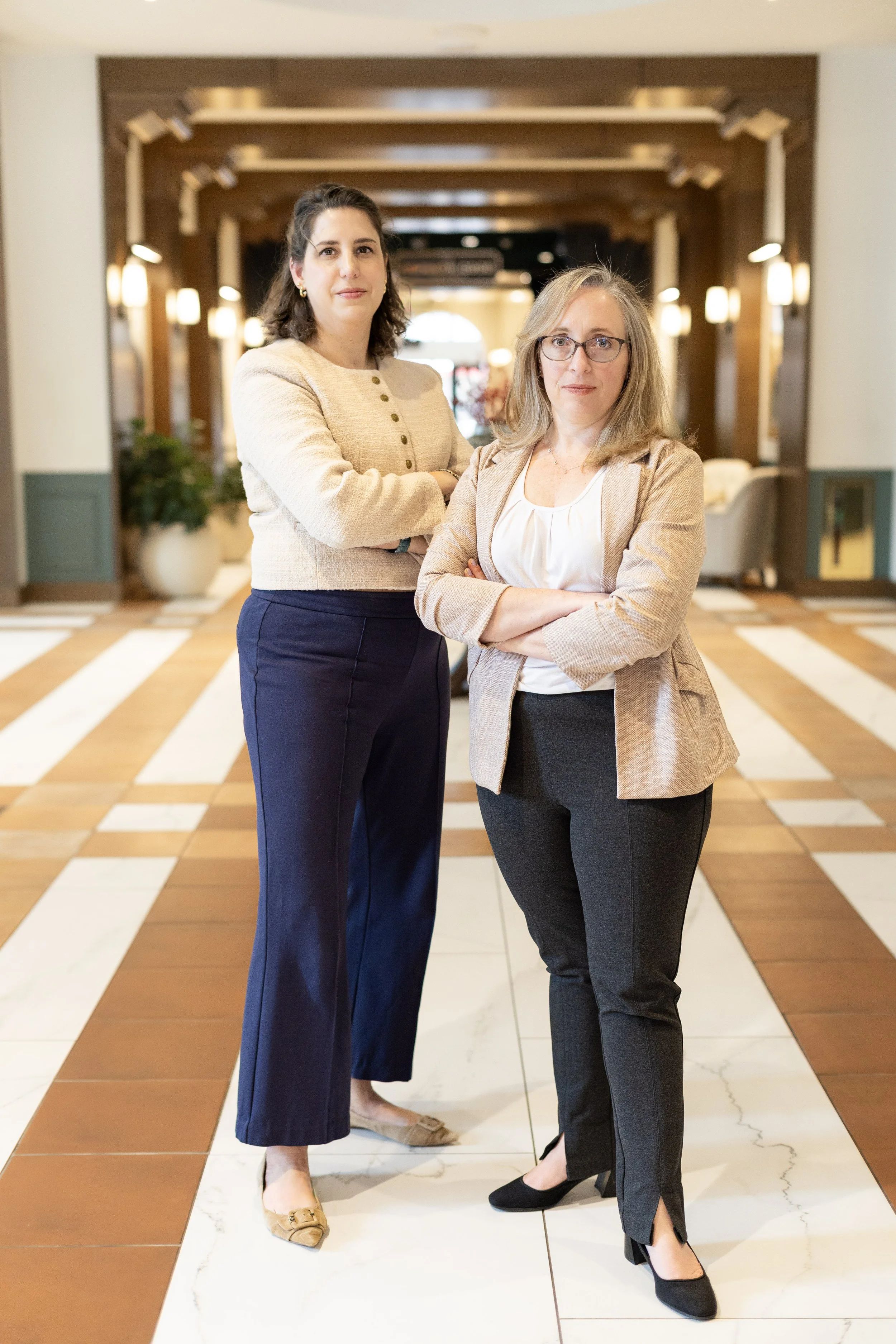 Two professional women standing in a hotel lobby. One woman with dark hair and a beige sweater, the other with gray hair and glasses, wearing a beige blazer.