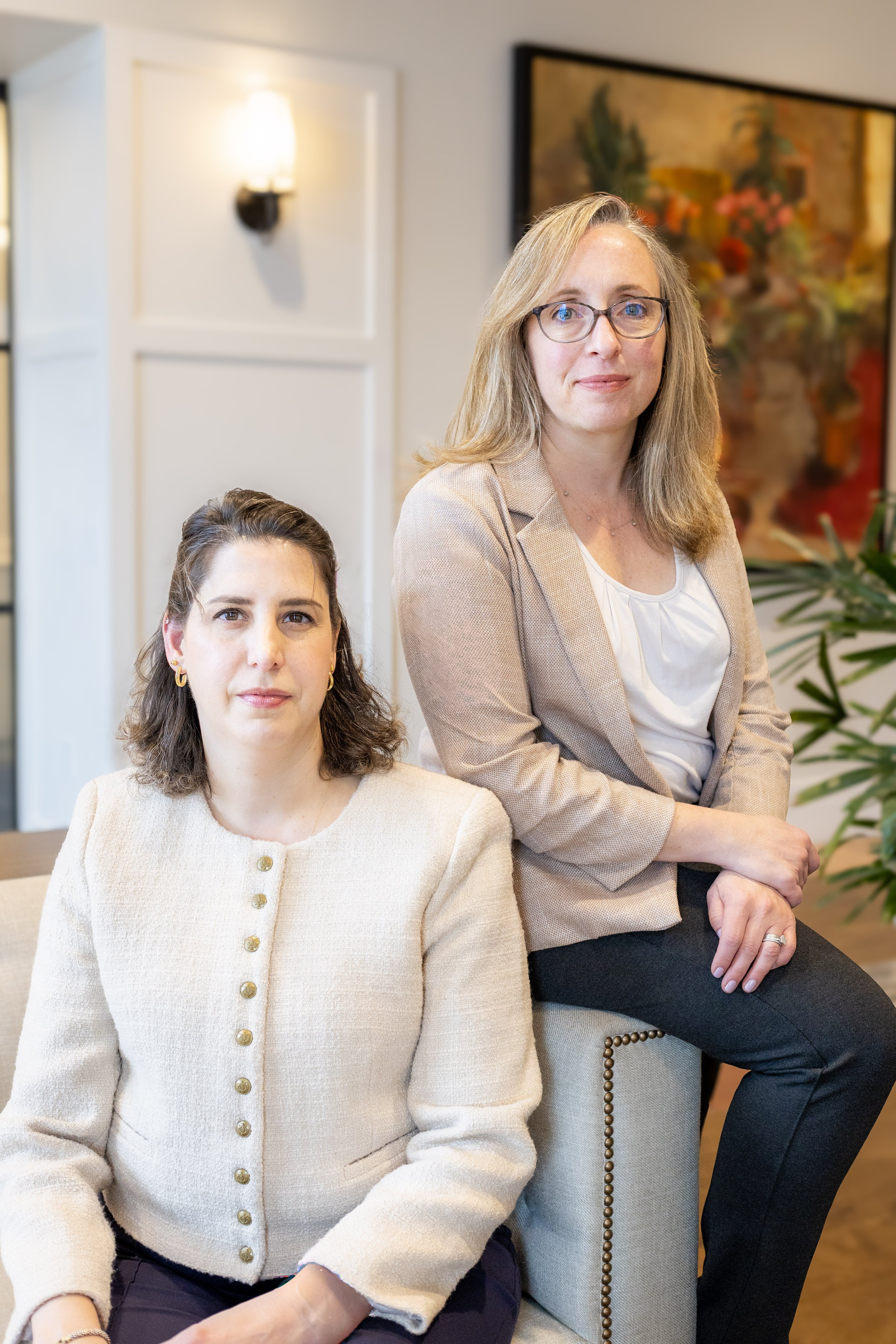 Two women in professional attire in an office setting, sitting near a wall with abstract art and a plant in the background.