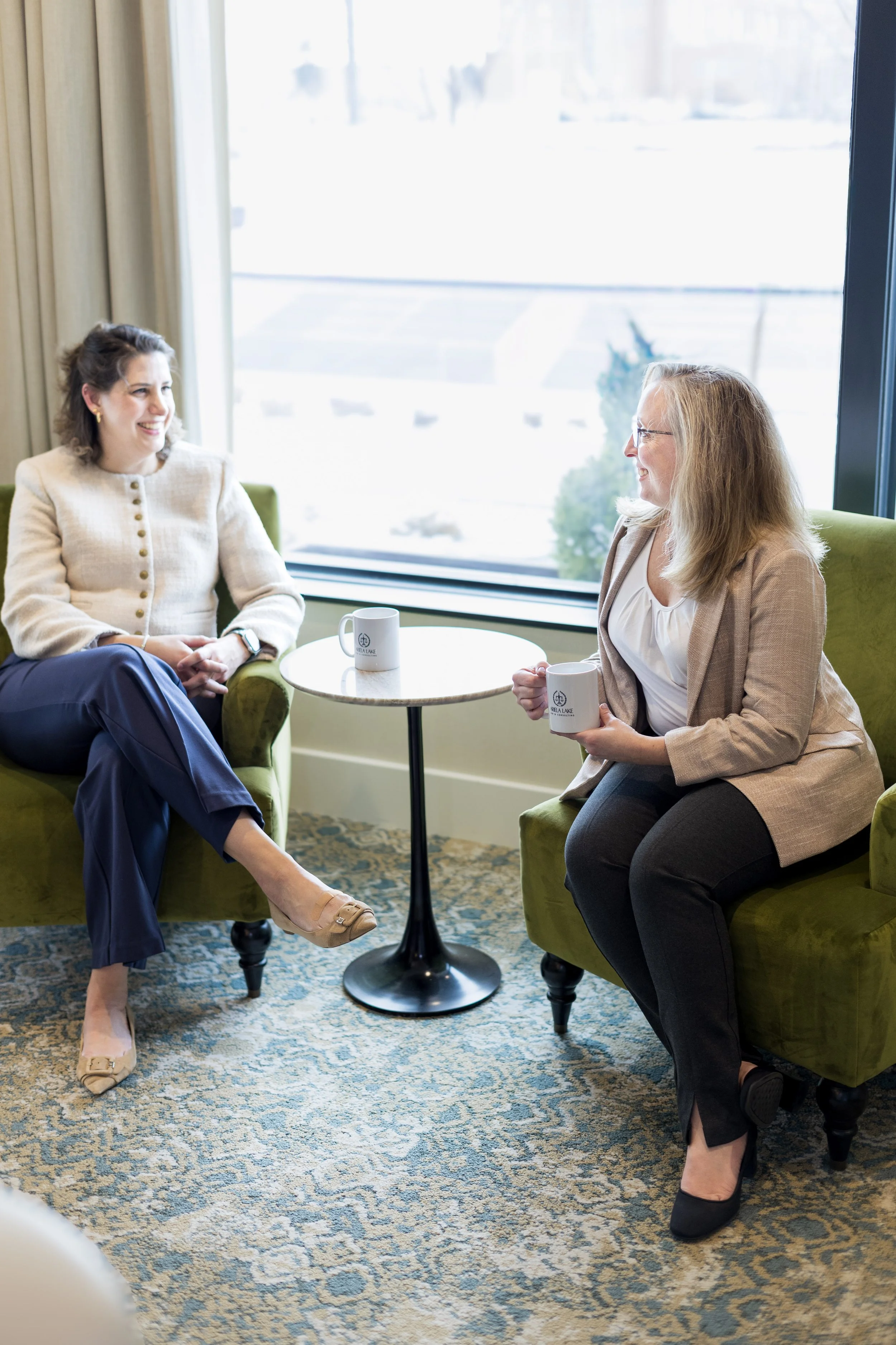 Two women engaging in a conversation while sitting on green armchairs near a window, each holding a white coffee mug with a logo, in a well-lit room with a patterned rug.