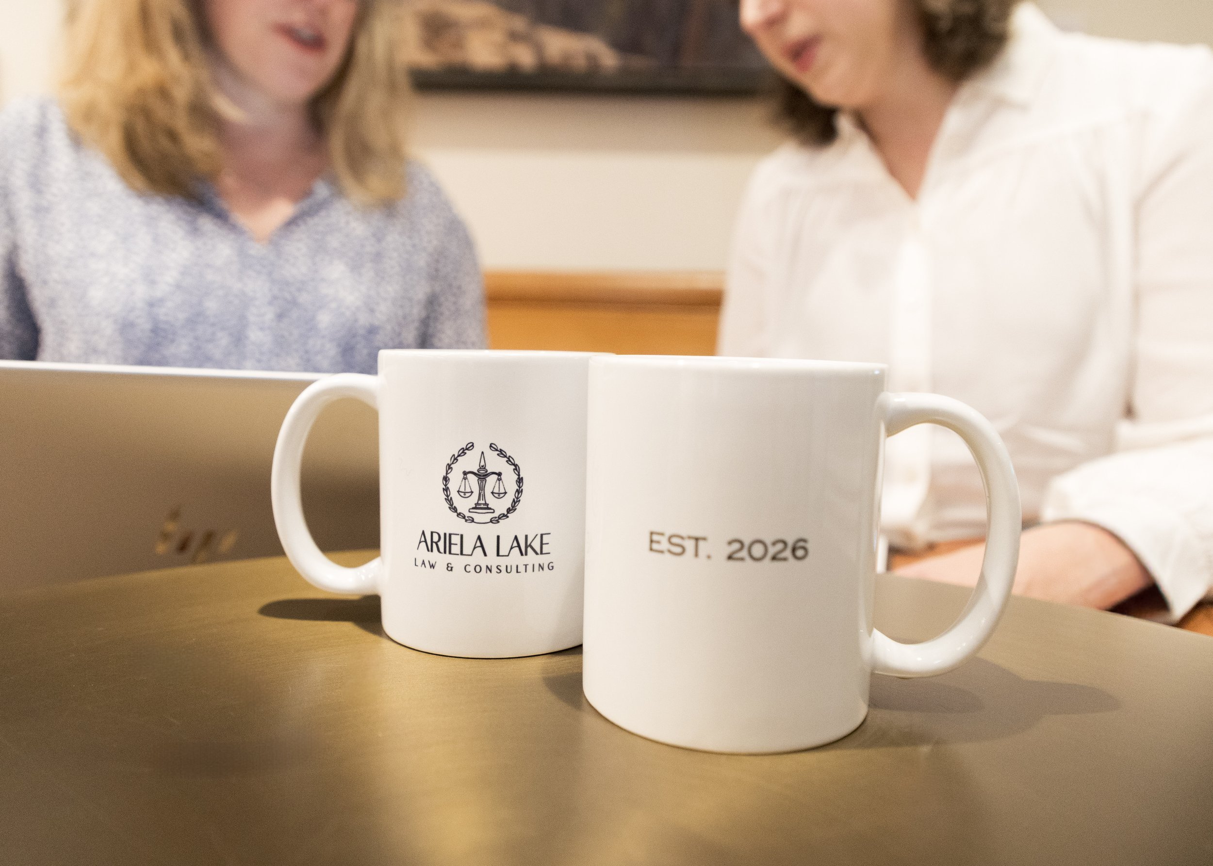 Two white coffee mugs on a wooden table with people in the background. One mug features the logo and name 'Ariela Lake Law & Consulting.' The other mug has 'EST. 2026' written on it.