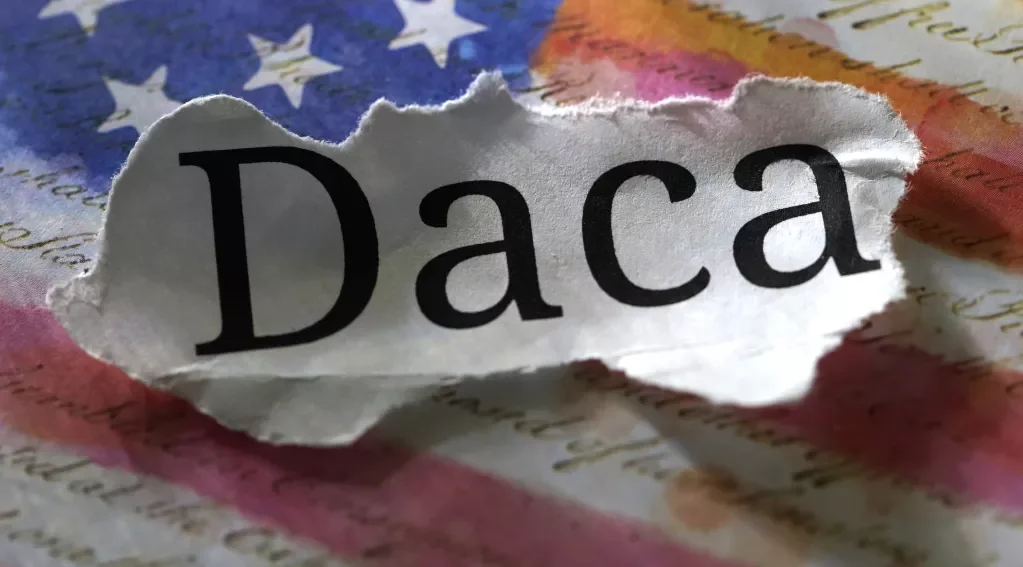 A torn piece of paper with the word 'Daca' printed on it, placed on a background resembling the American flag.