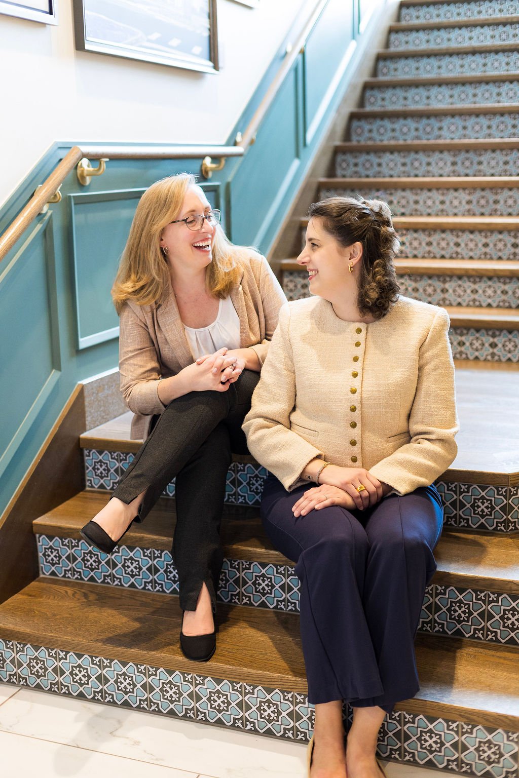 Two women sitting on stairs, smiling and looking at each other, in an interior space with patterned tiles on the staircase.