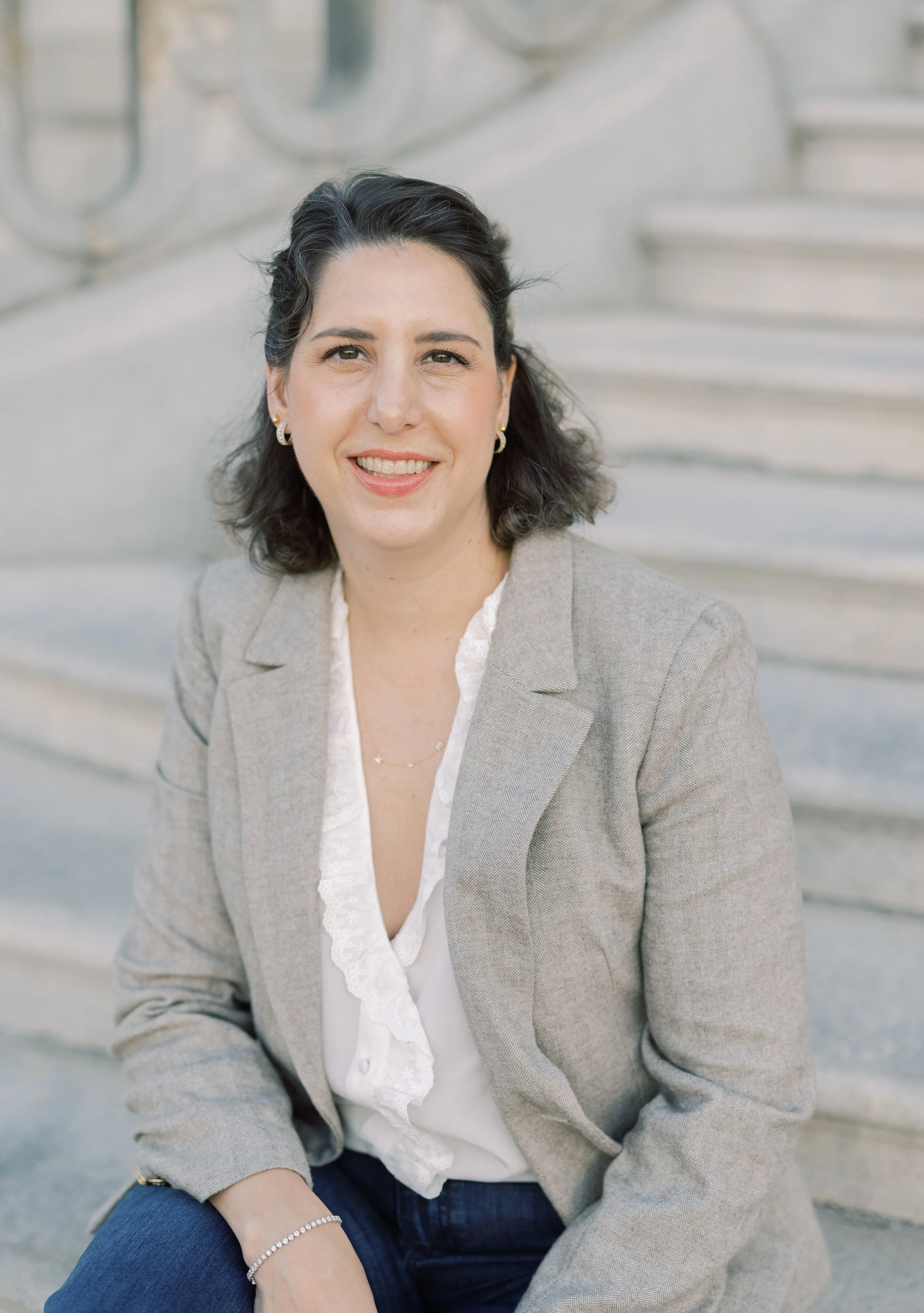 A woman with dark, curly hair, dressed in a light gray blazer, white blouse with ruffles, and jeans, sitting on outdoor steps and smiling at the camera.
