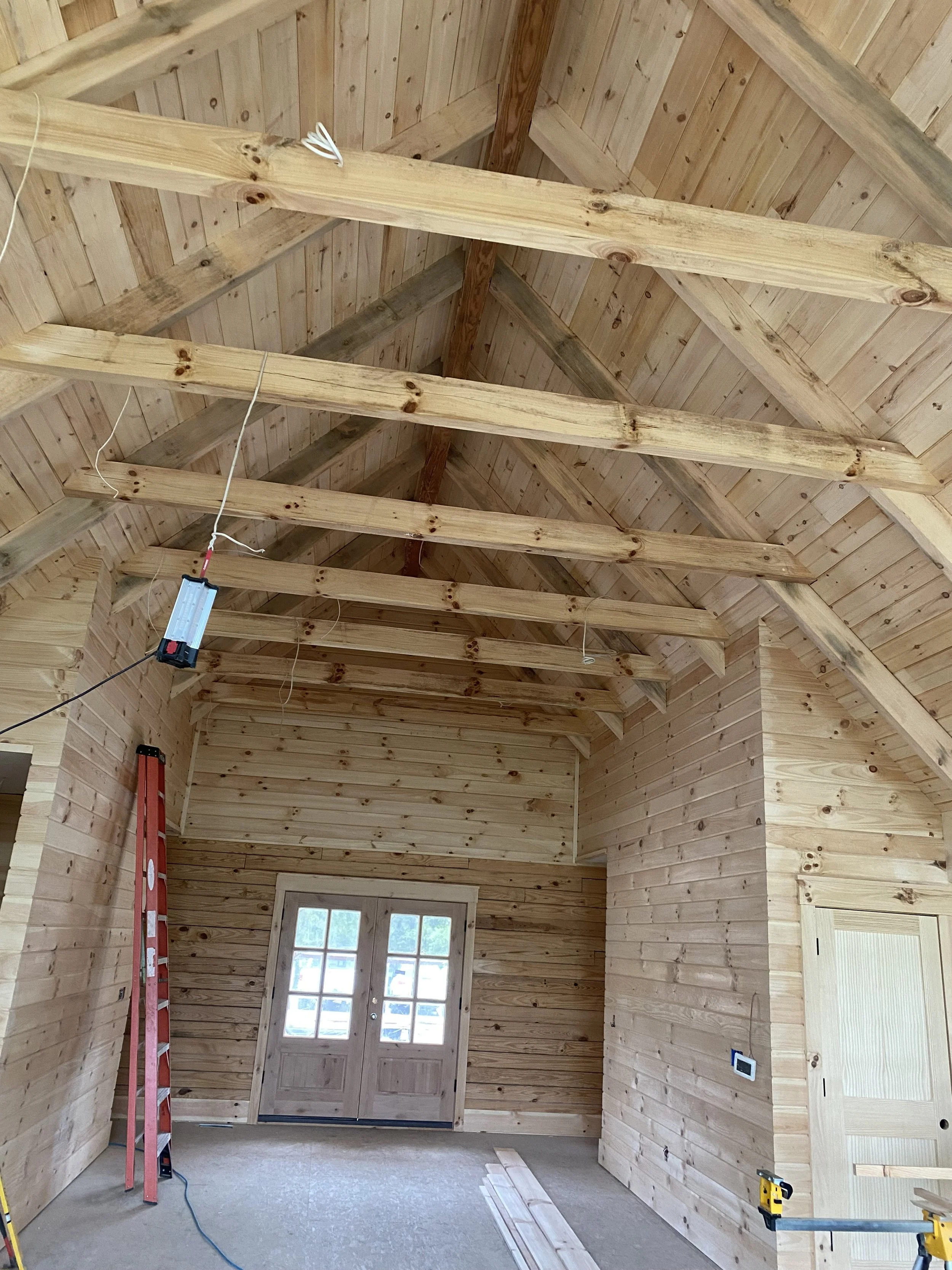 Interior of a wooden cabin under construction with exposed roof beams, ladder, and a door with small windows.