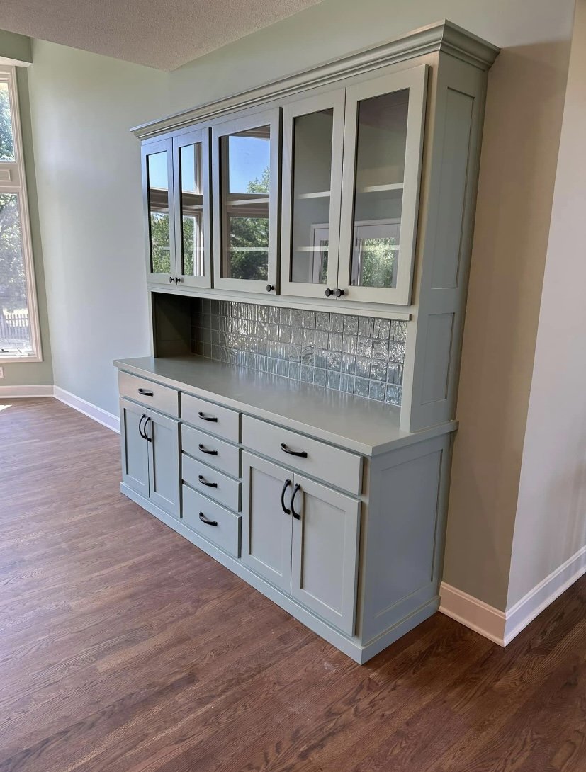 Light gray kitchen hutch with glass doors on the top section, drawers and cabinets on the bottom, set against a beige wall in a room with hardwood floors.