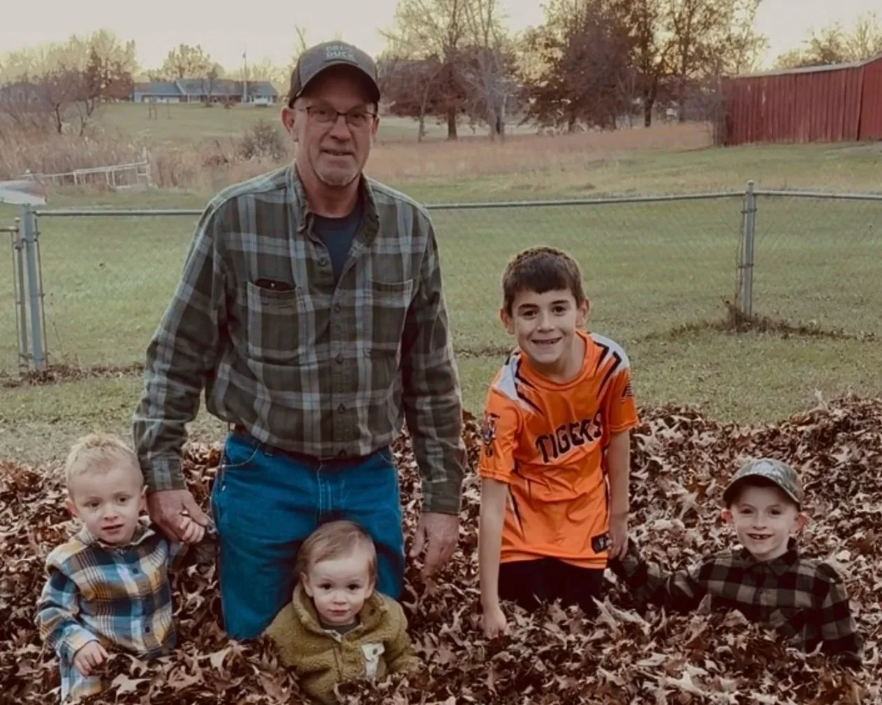 A man and four children in a backyard surrounded by fallen autumn leaves during sunset.
