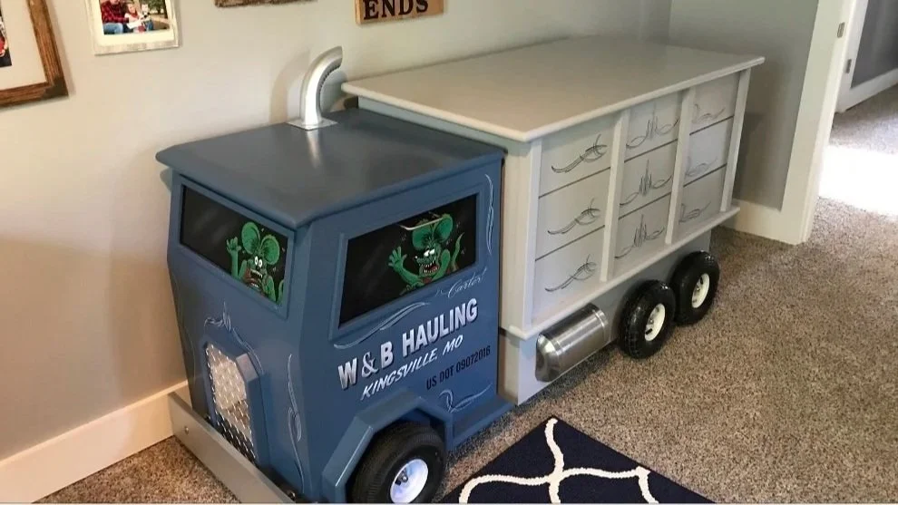 A children's playhouse or furniture piece designed to look like a moving truck, with cartoon characters inside the windows and decorative lettering, situated on carpeted flooring near a wall with family photos.