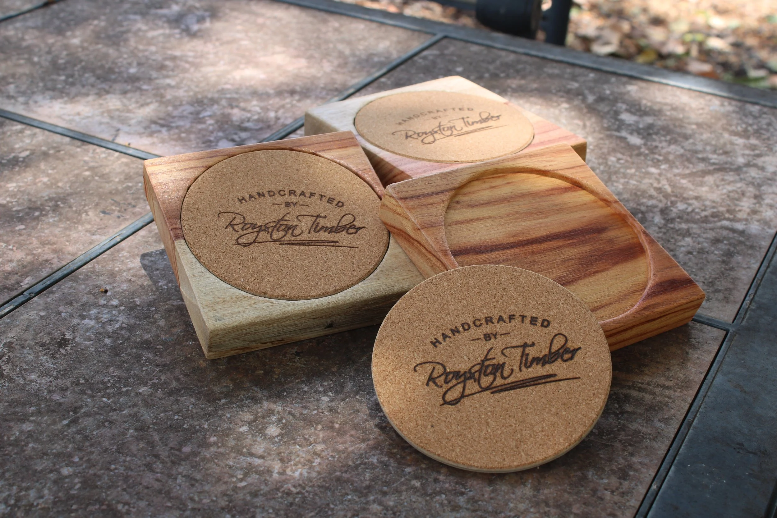 Wooden coasters with the text 'Handcrafted by Rayston Timber' on cork surfaces, placed on a textured outdoor table.