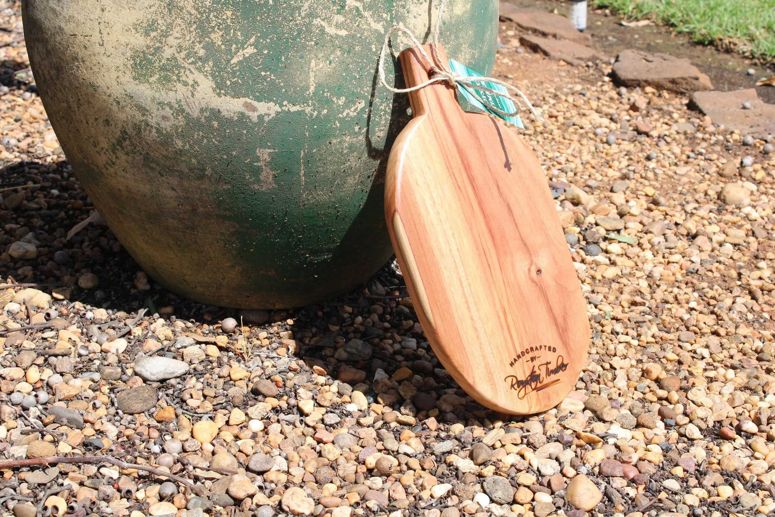A wooden cutting board leaning against a weathered green ceramic pot on a gravel surface.