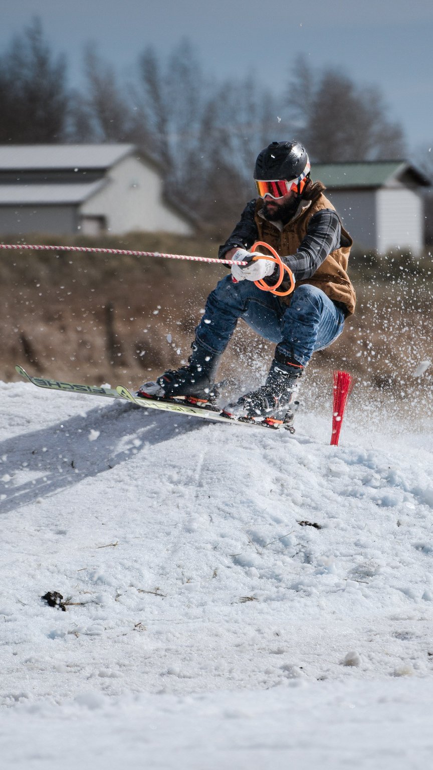 A man skijoring, wearing a black helmet, orange goggles, brown vest, and jeans is skiing on snow, holding a red rope, with a red marker on the snow, and houses and trees in the background.