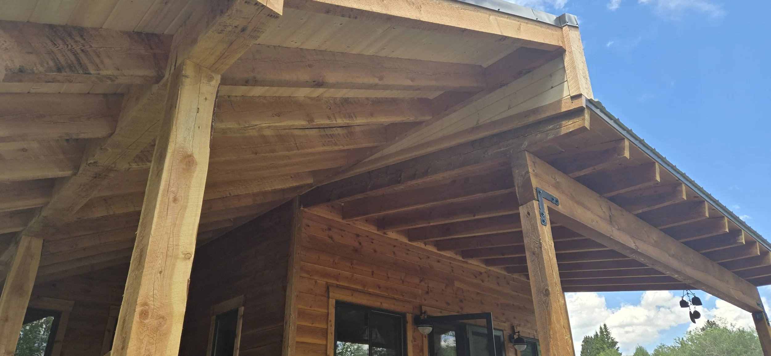 Close-up view of the wooden porch and roof extension of a house, with visible support beams, under a partly cloudy sky, and trees in the background.