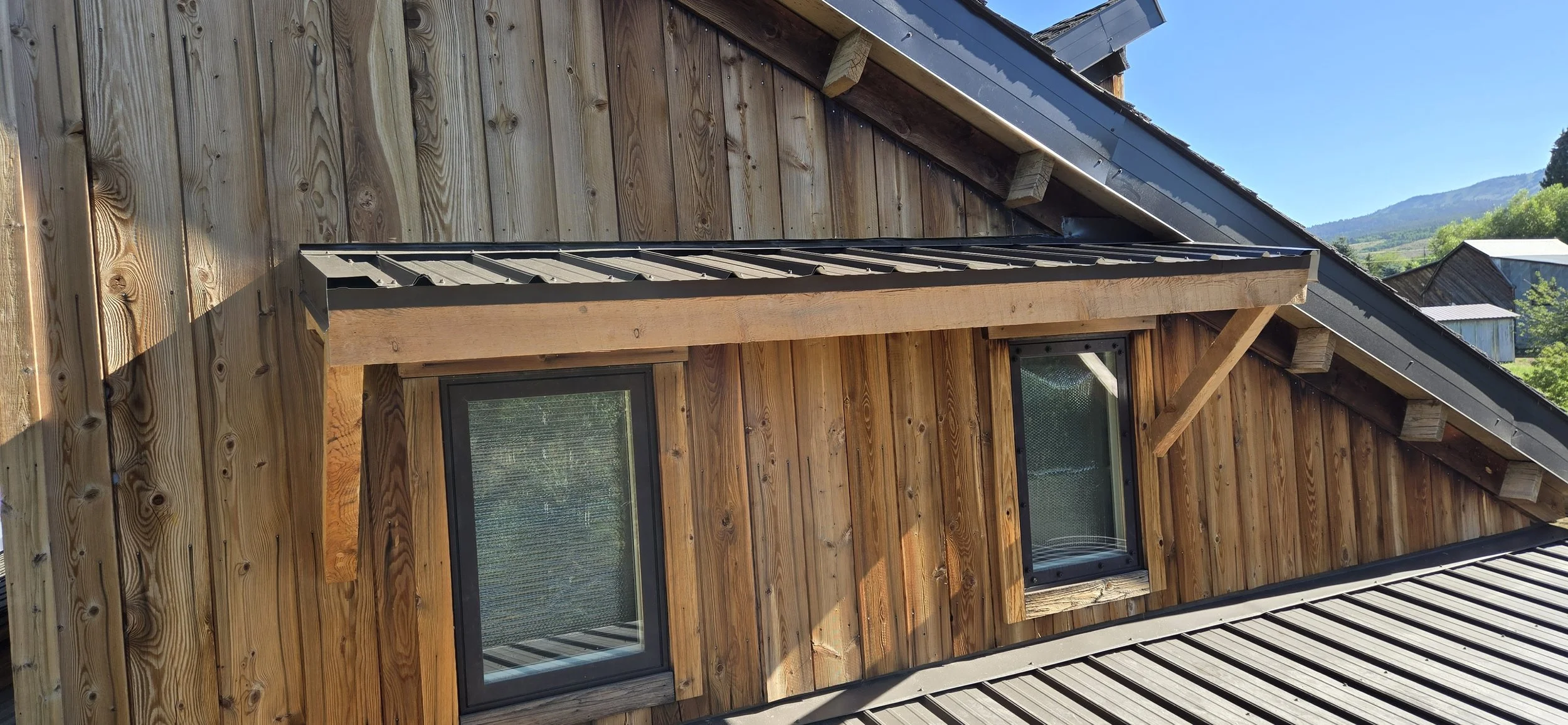 Close-up of a wooden house with two black-framed windows and a metal roof in a scenic, mountainous area with clear blue sky.