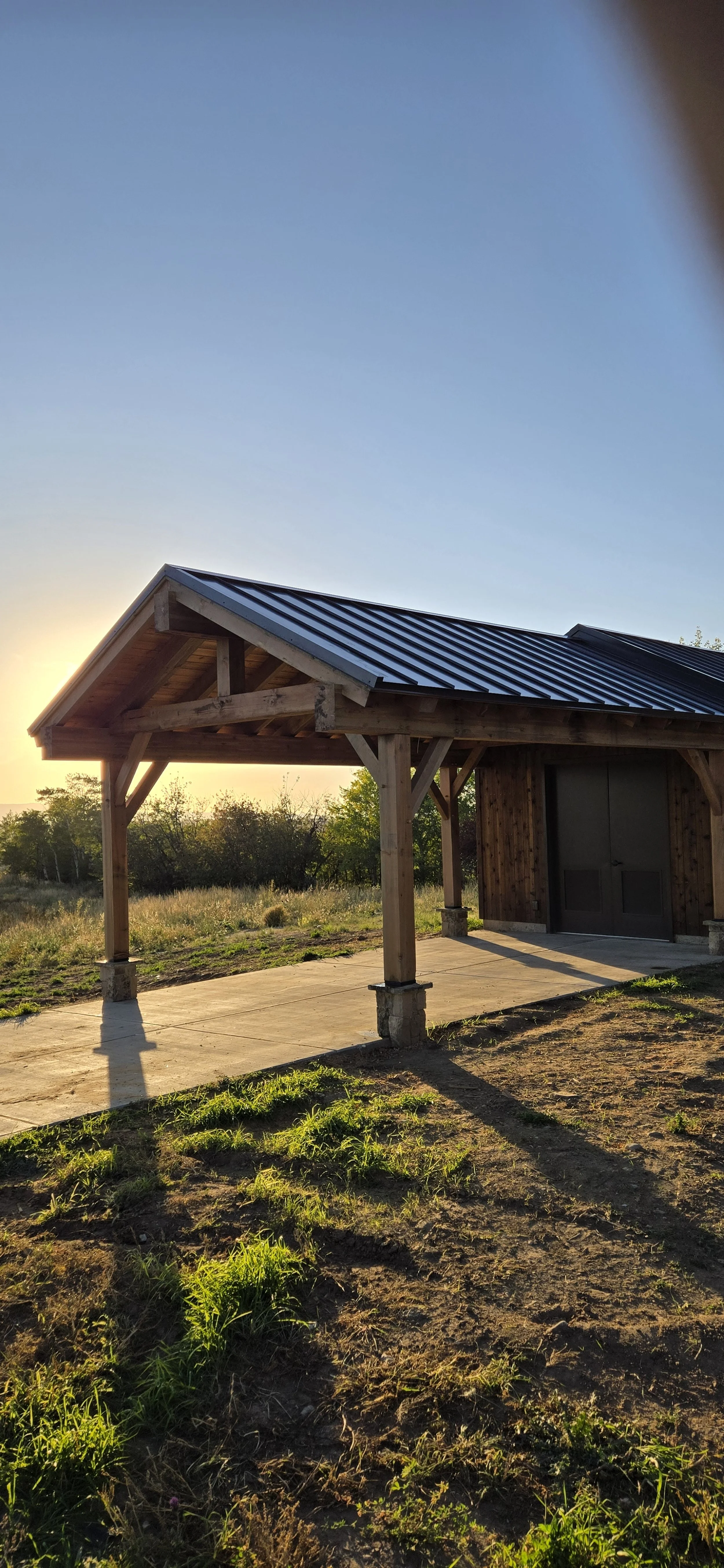 A wooden structure with a metal roof standing on a concrete pad, surrounded by grass and trees, with the sun setting in the background.