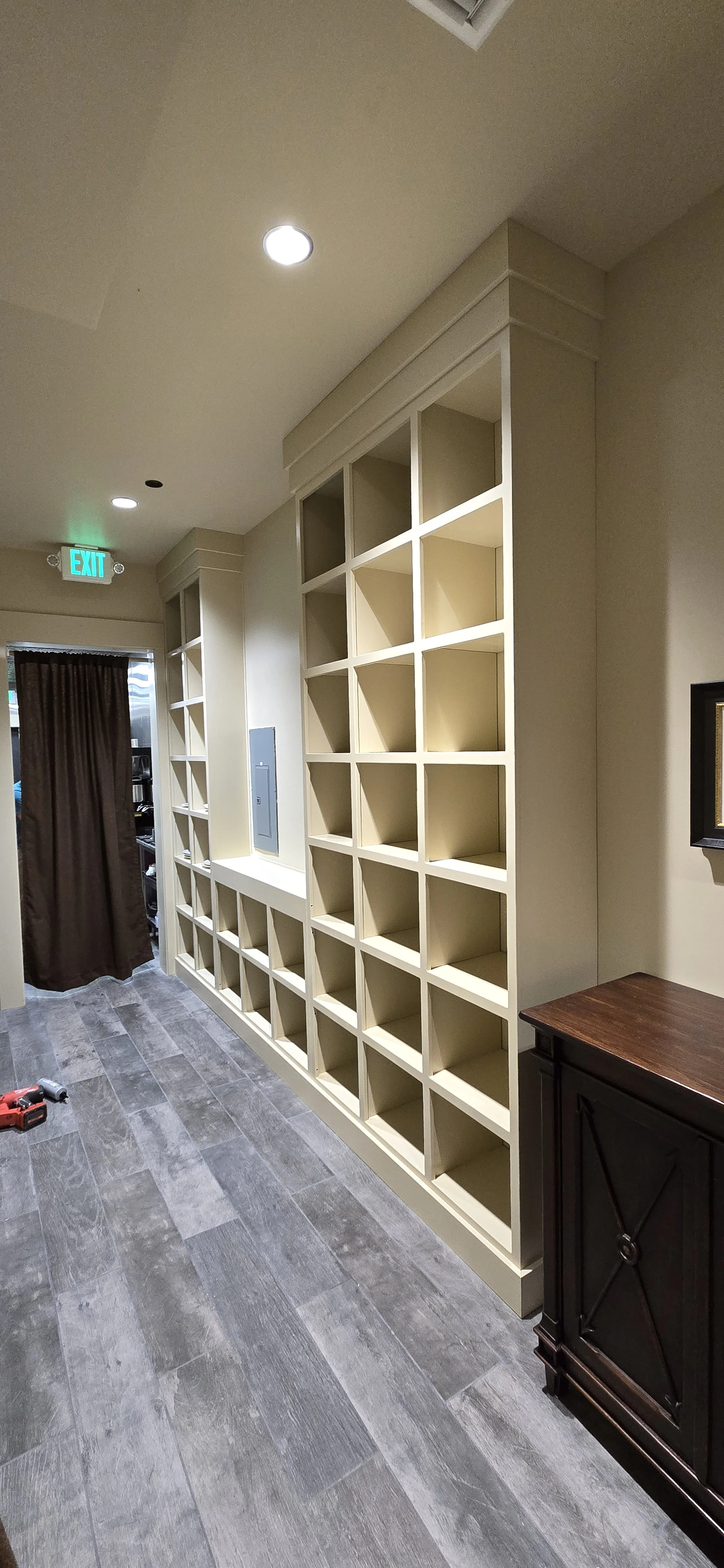 Empty built-in wall shelves in a room with gray wood-like flooring, a black cabinet, and a doorway with a curtain. Recessed ceiling lights and a green exit sign are visible.
