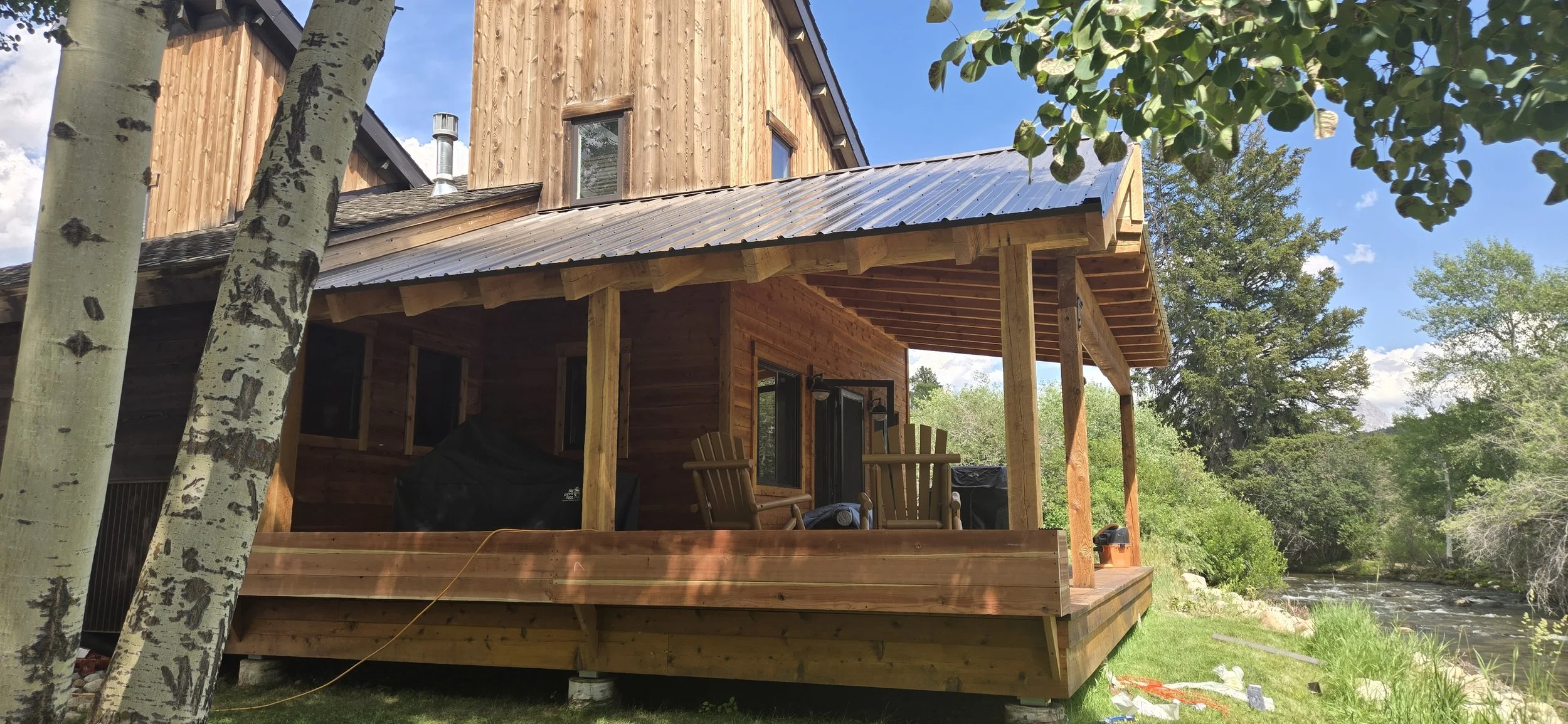 A wooden house with a covered porch and metal roof, situated beside a stream, surrounded by trees and greenery, with outdoor chairs on the porch.