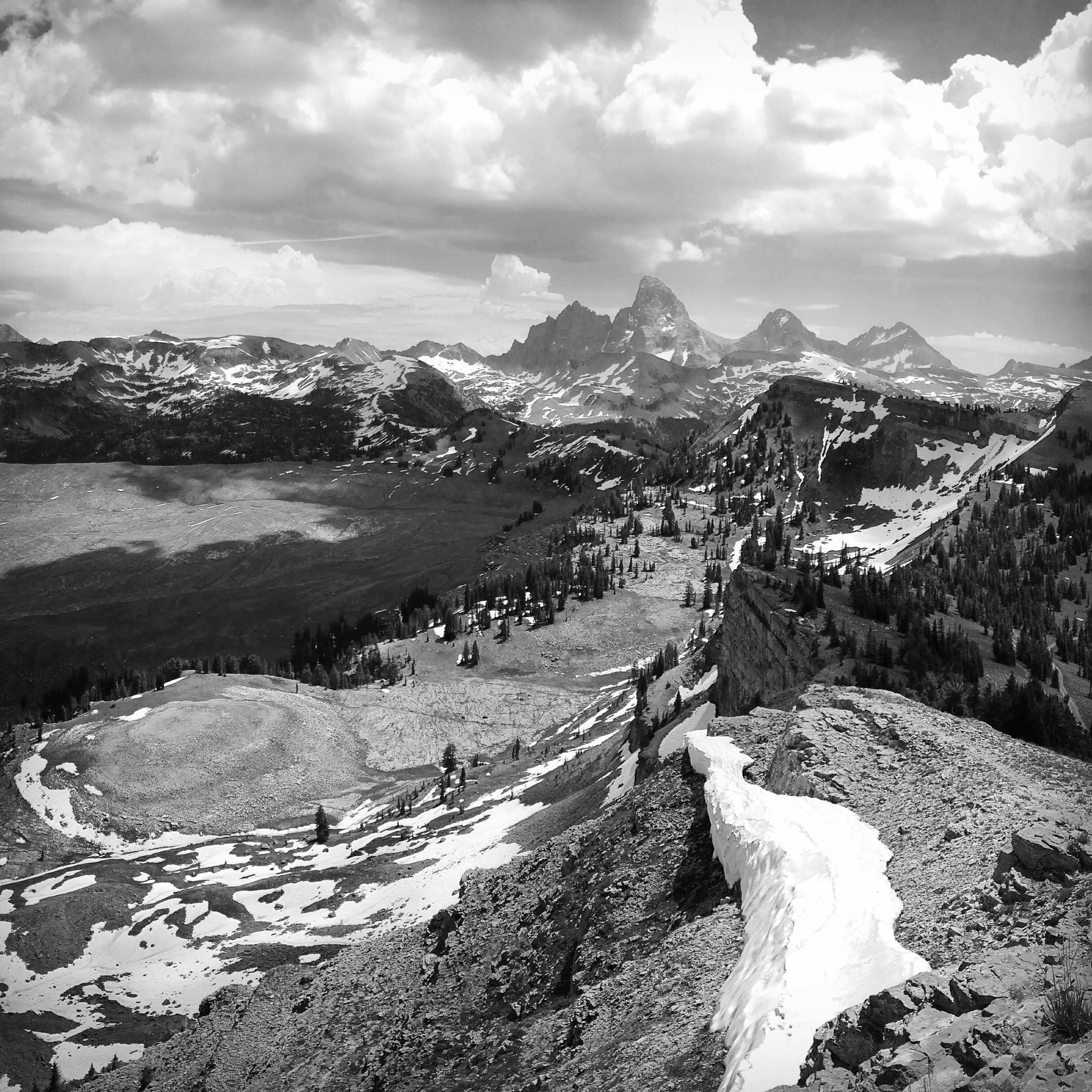 Black and white photo of a mountain landscape with snow patches, rocky ridges, and distant peaks, including a prominent sharp mountain summit.