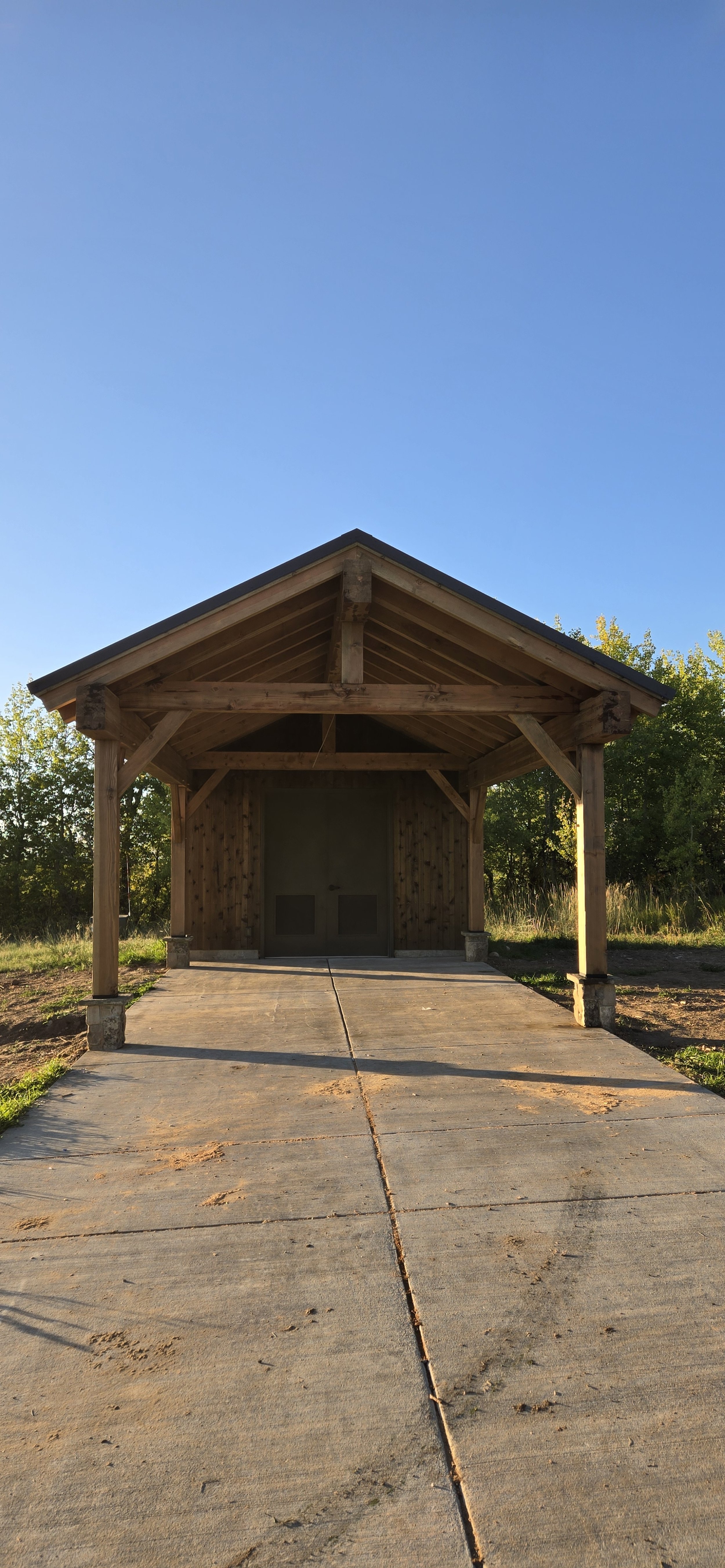 A wooden pavilion with a pitched roof and sliding doors, situated outdoors on a concrete path with grass and trees in the background under a clear blue sky.