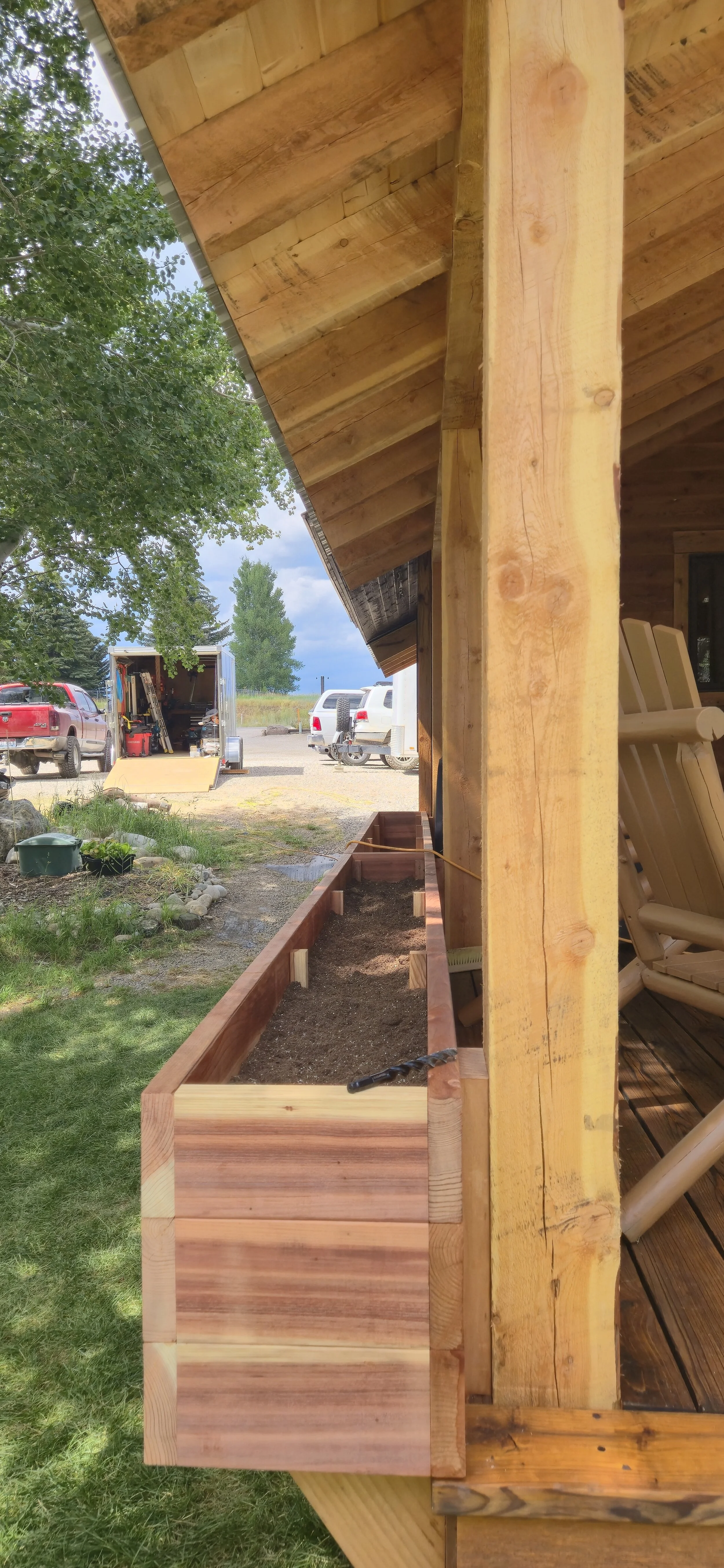 Side view of a wooden porch or deck under construction, with a planters box in the foreground and a trailer with tools and equipment in the background, in a rural setting with trees and colorful vehicles.