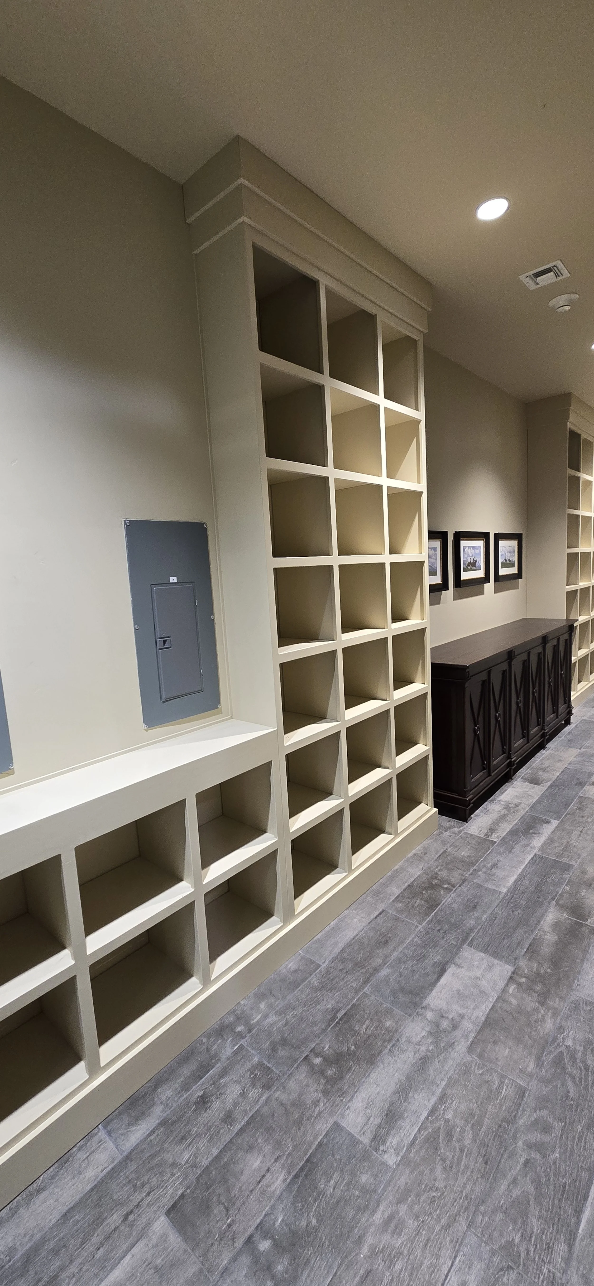 Empty shelving units against a beige wall in a room with gray hardwood flooring and framed pictures, with an electrical panel on the wall.