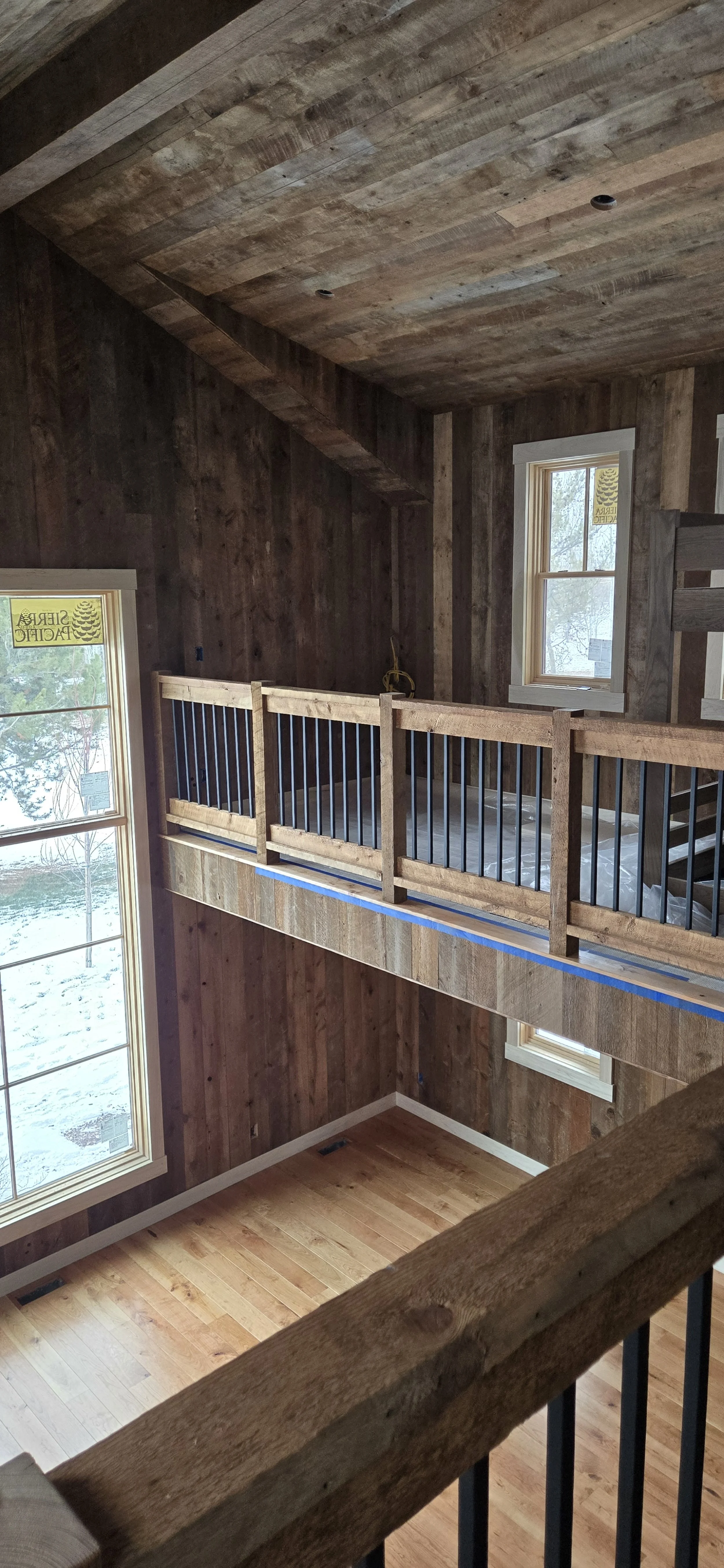 Interior view of a cabin under construction with wood-paneled walls and ceiling, a wooden railing on a loft, two windows letting in natural light, and hardwood flooring.