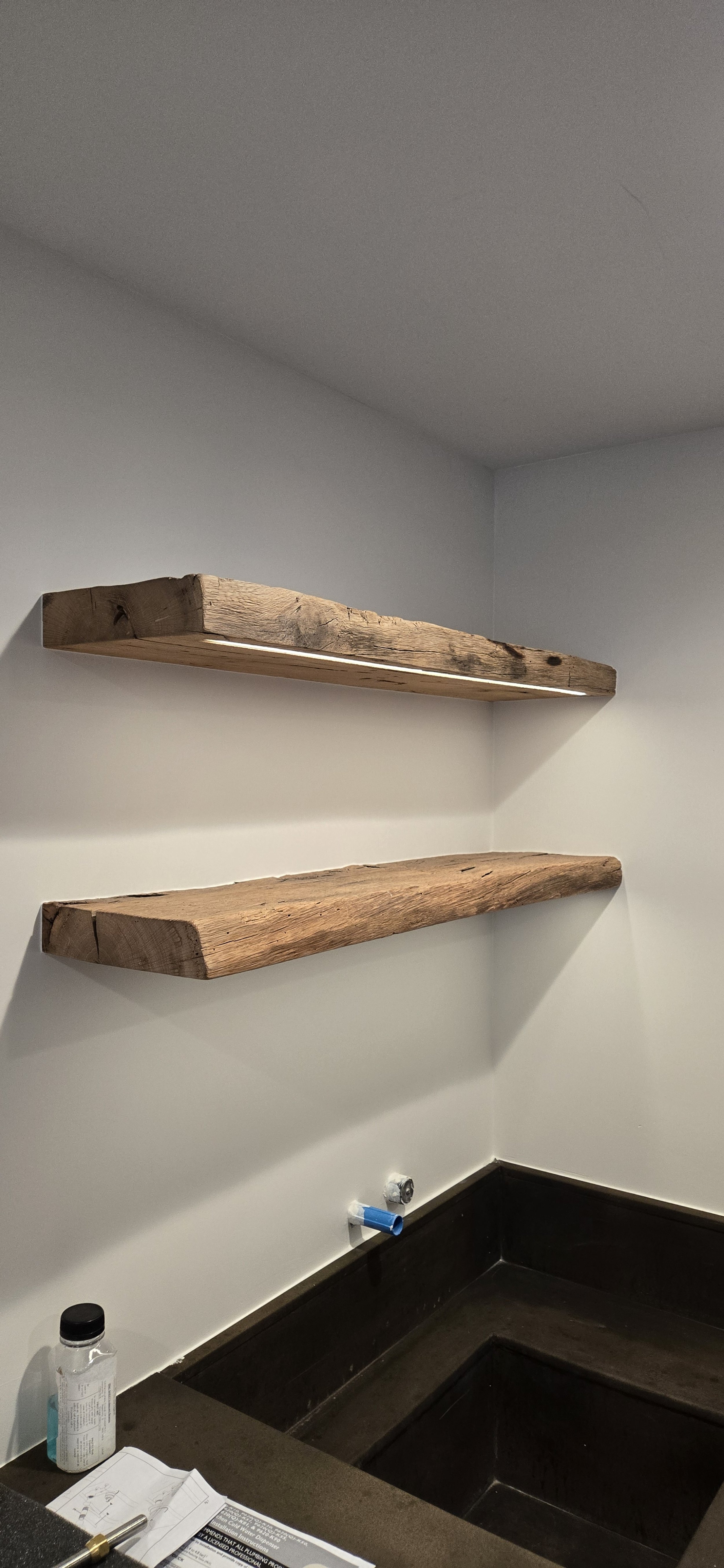 Two rustic wooden shelves mounted on a white wall above a dark countertop with a sink cutout. Items on the counter include a bottle, some papers, and a pen, indicating a kitchen or utility area.