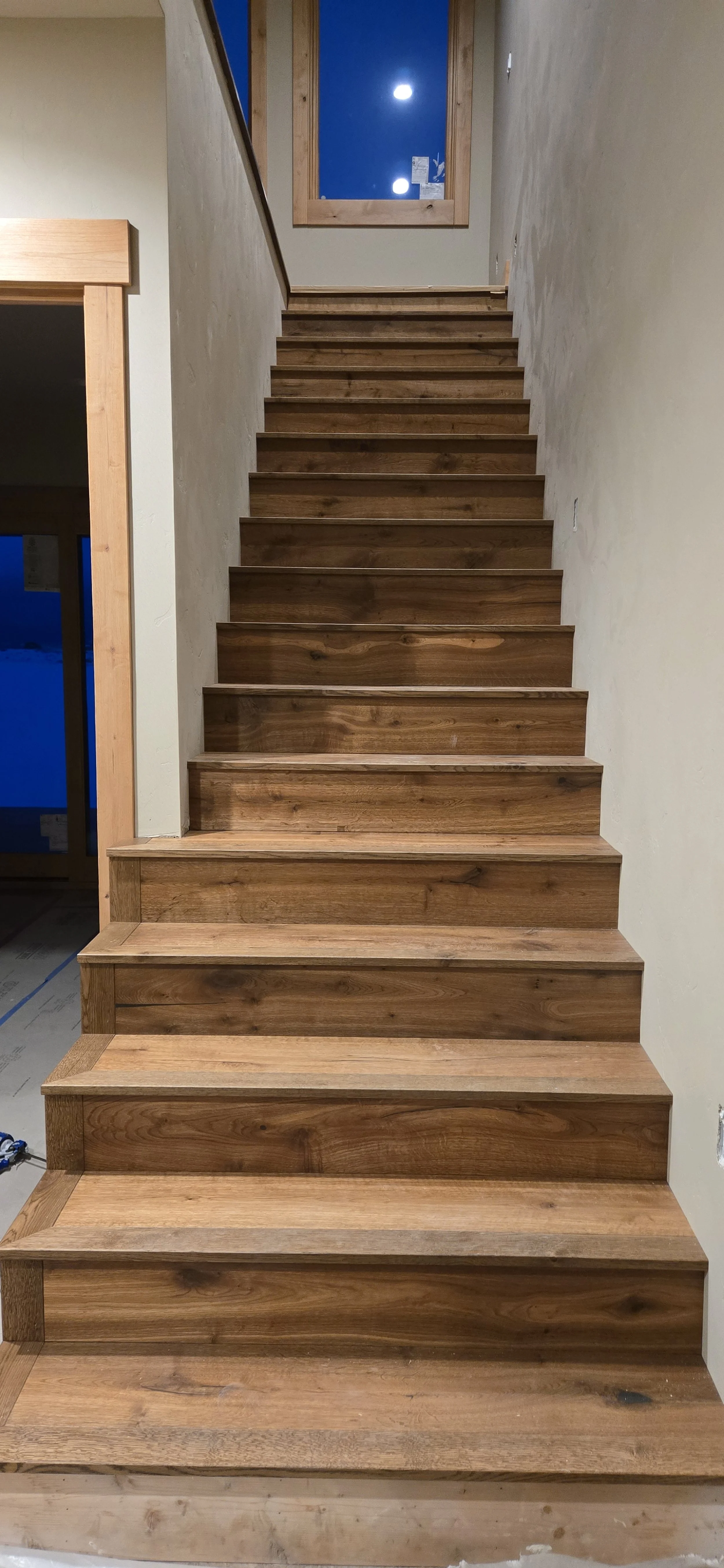 Wooden staircase inside a house, leading upward toward a window at the landing. The stairs are made of natural wood, and part of the interior wall is visible with an unfinished or textured surface.