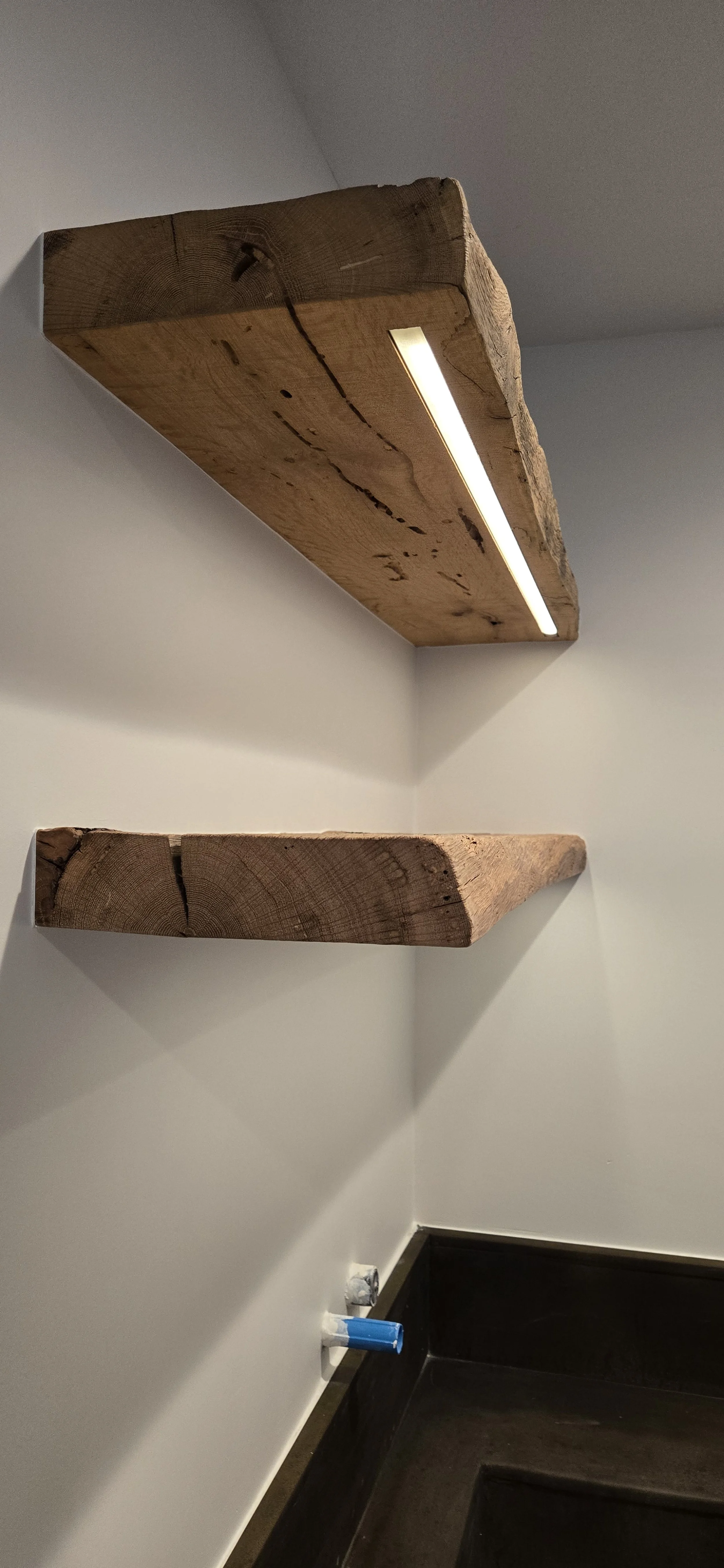 Two wooden shelves attached to a white wall, one with an integrated light strip, and a blue pipe protruding from the wall near the floor.
