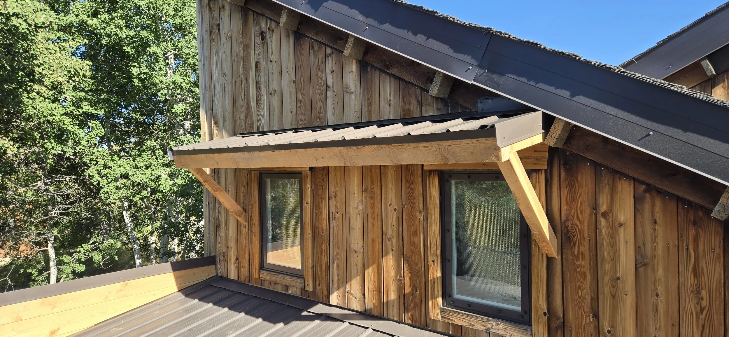 Close-up of the exterior of a wooden house with a dormer window and a sloped roof, surrounded by lush green trees.