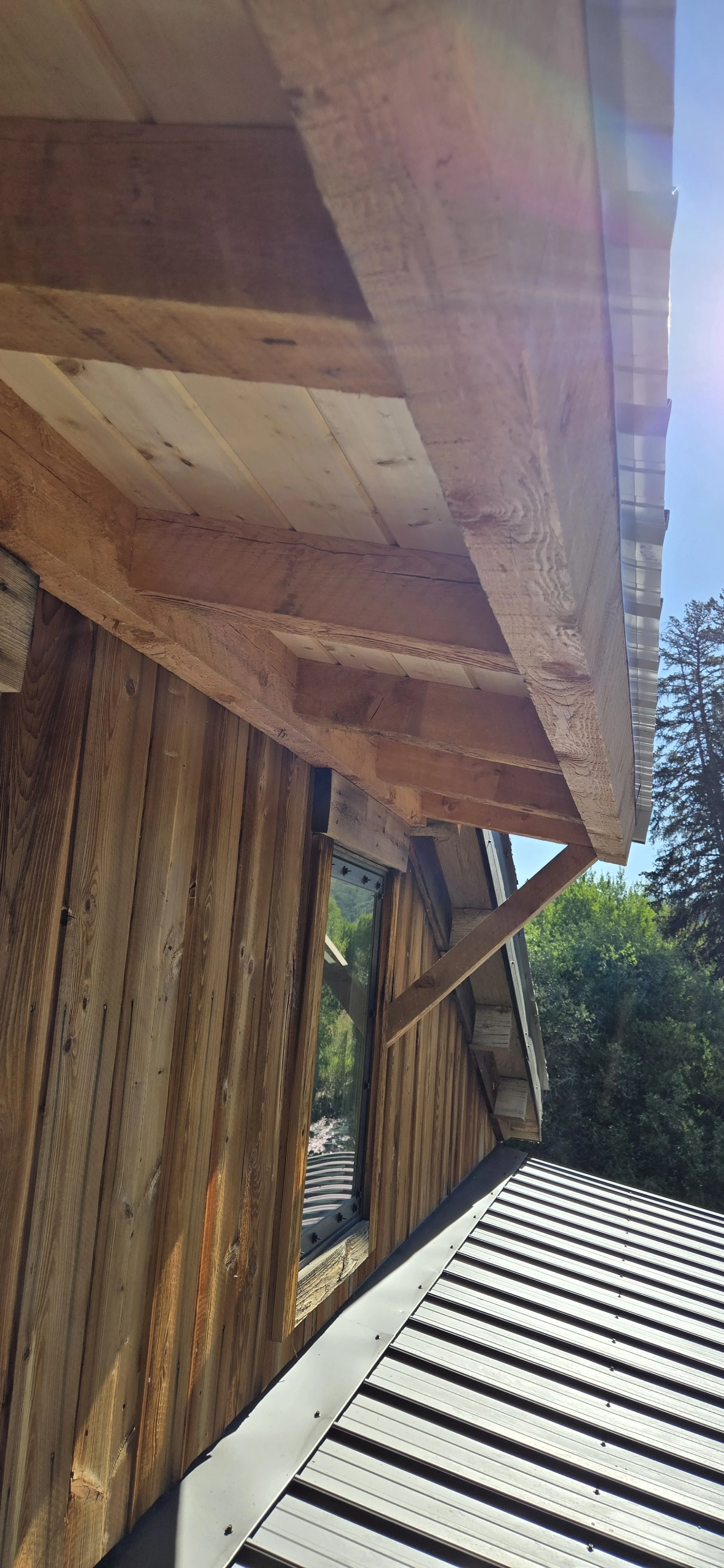Close-up of a wooden building exterior, featuring a window and a metal roof, with trees in the background and sunlight creating lens flare.