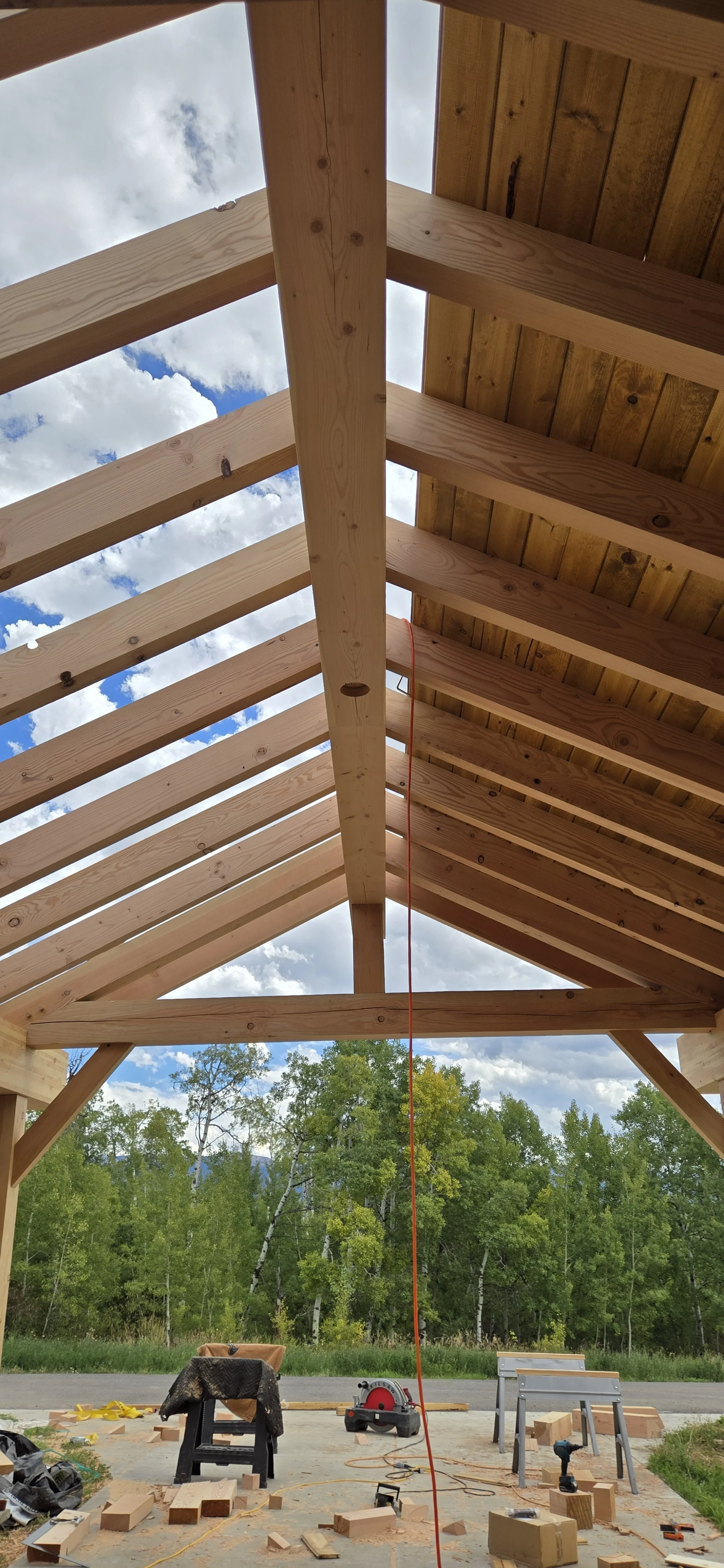 View looking up at a wooden roof structure under construction, with blue sky and clouds visible through the open sections, and trees in the background.