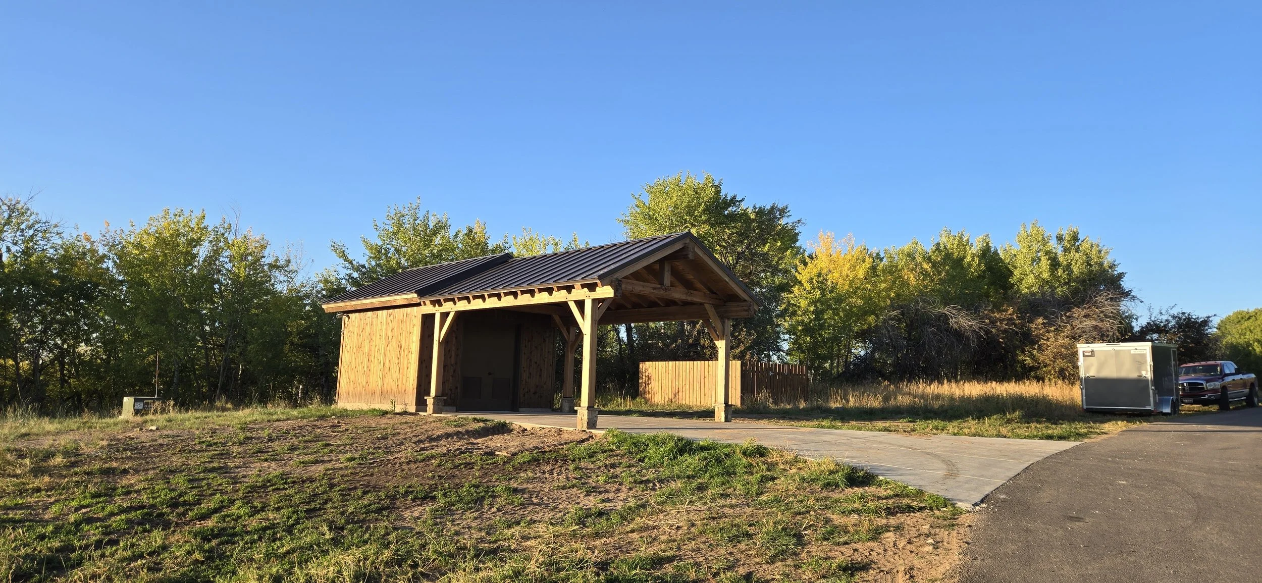 A small wooden shed with a metal roof, surrounded by trees, with a paved driveway and a truck with a trailer attached, under a clear blue sky.