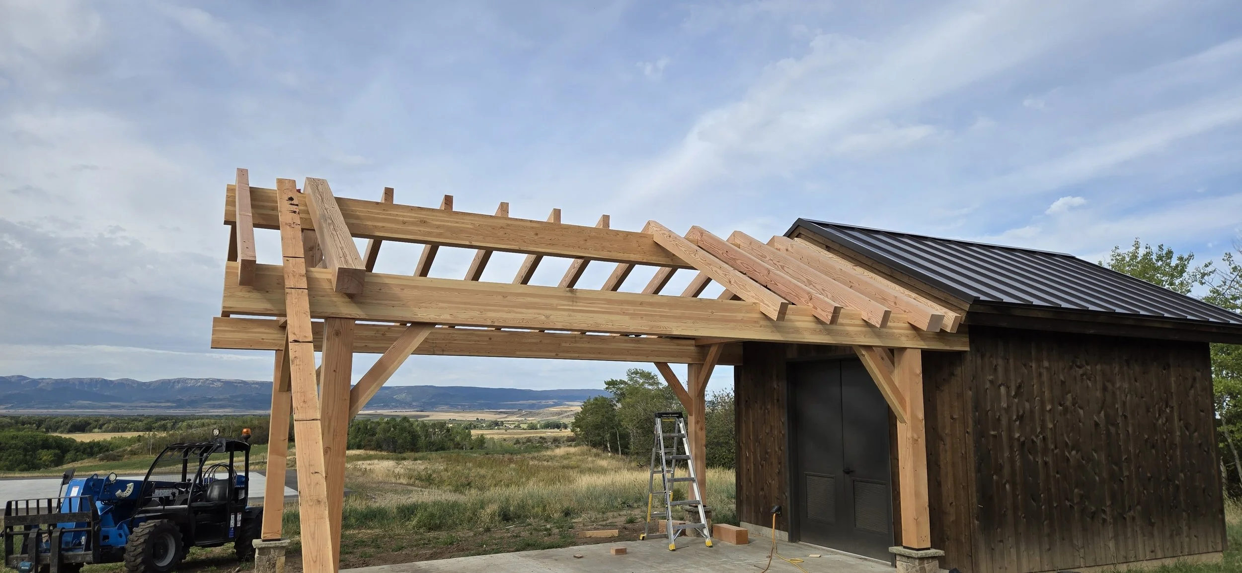 Construction of a wooden shelter with a metal roof, showing the framing in progress, set against a rural landscape with mountains in the background.