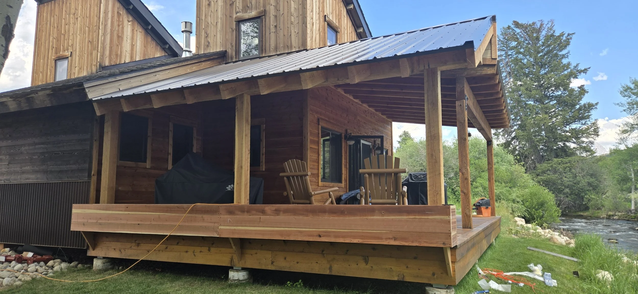 A wooden house with a porch overlooking a river, surrounded by green trees and grass, with construction tools and materials nearby.